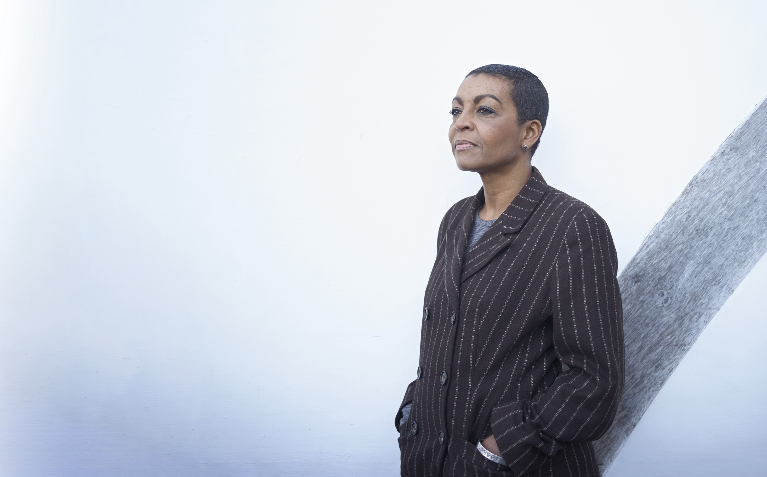 Adjoa Andoh A black woman with short cropped hair stands against a white wall wearing a dark pinstripe suite.