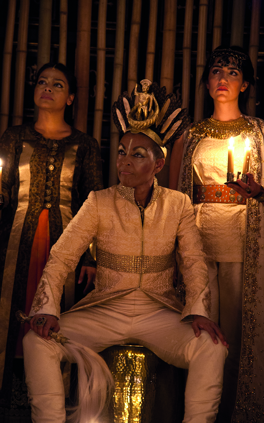 An actor in a white suit sits on a throne wearing a gold crown