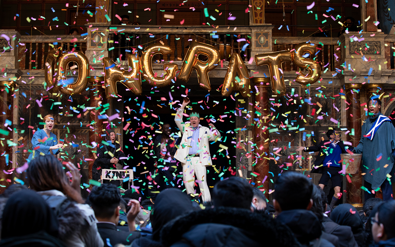 An actor has balloons spelling congrats above him while confetti showers the stage
