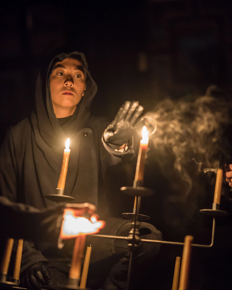 MACBETH A cloaked man raises his hand to a lit candle.