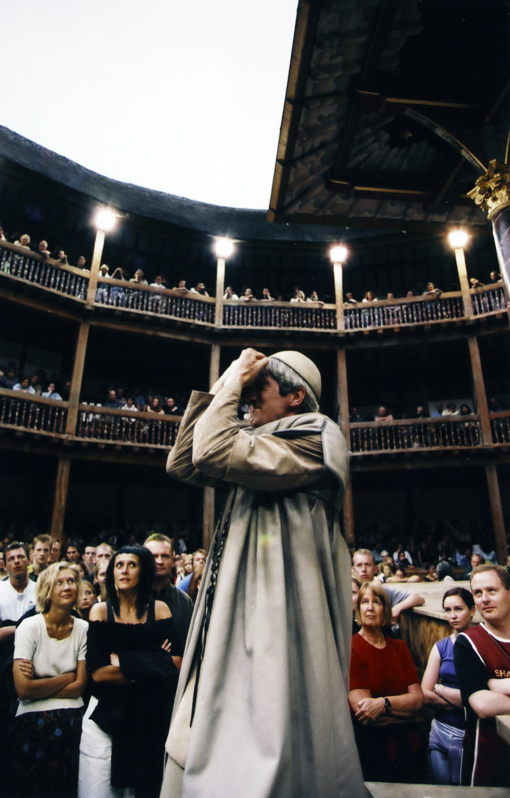 A man holding his hands up to his face stands in the Yard of the Globe Theatre, surrounded by audience members.
