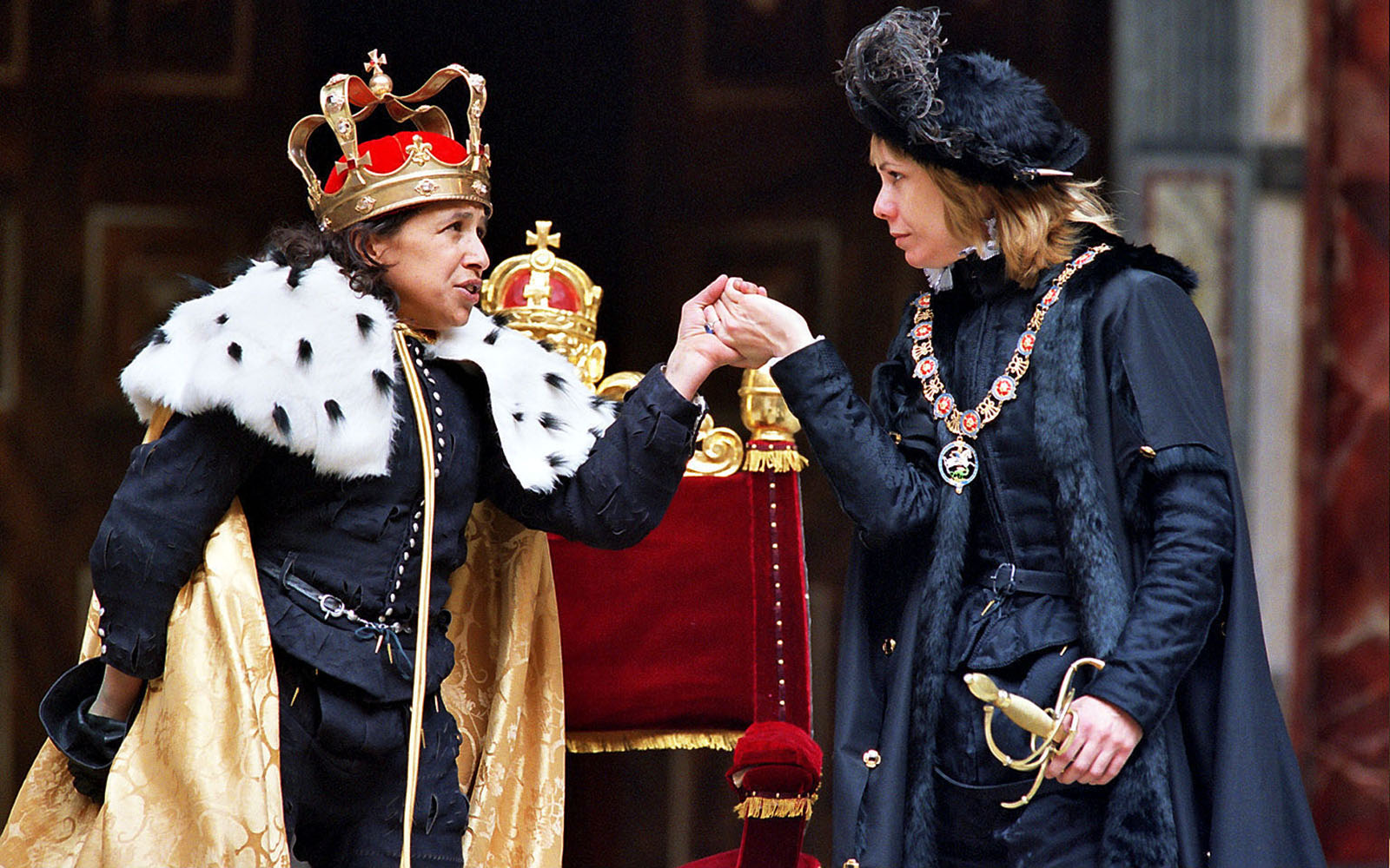An actor with a red crown and wearing a golden ermine cloak reaches to kiss the hand of another woman wearing black traditional Elizabethan dress.