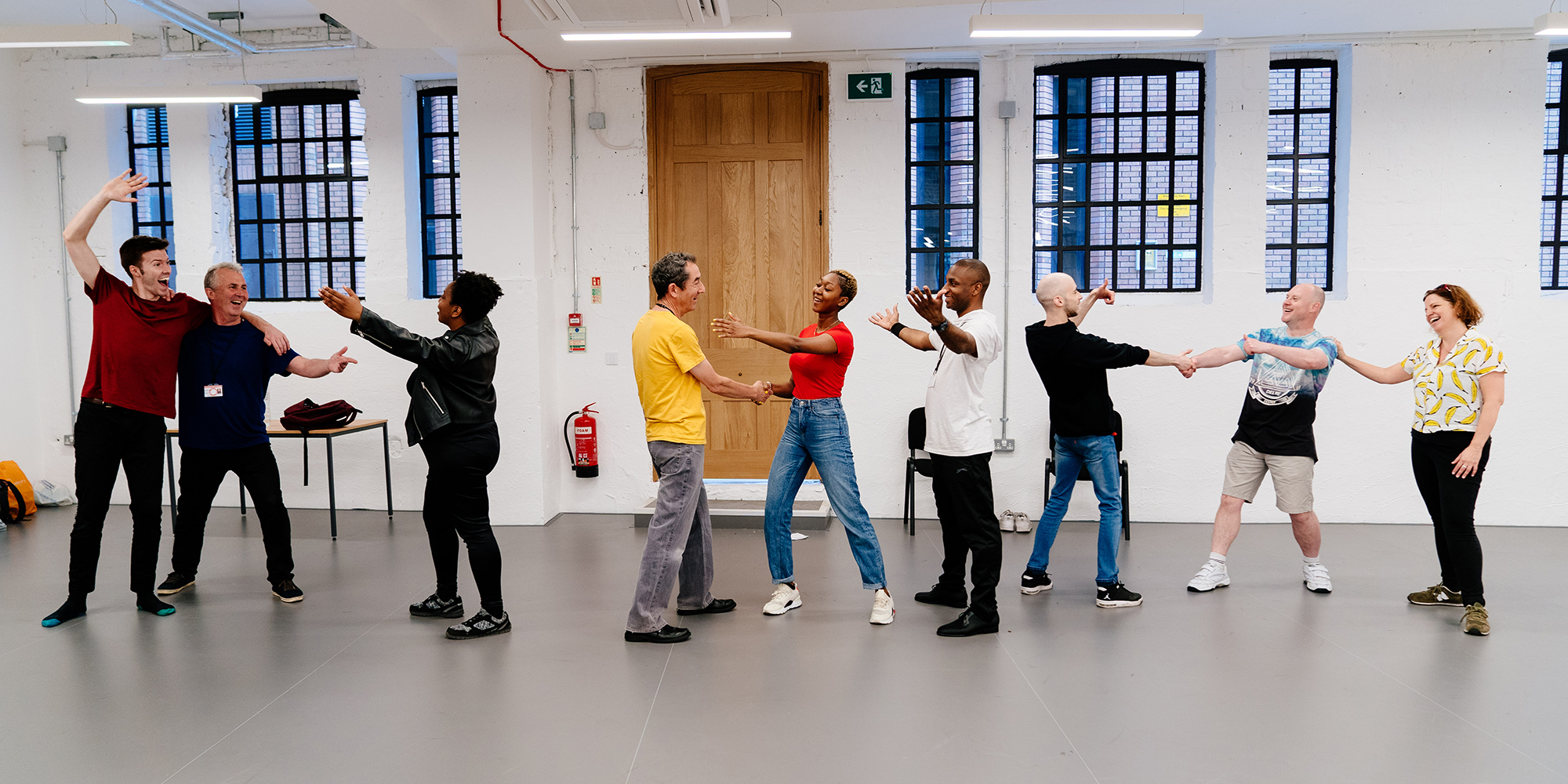 London Bubble The Windsor Locals Merry Wives of Windsor Shakespeare's Globe 2019 A chain of people stand in a row in a rehearsal room, arms outstretched and smiling at each other.