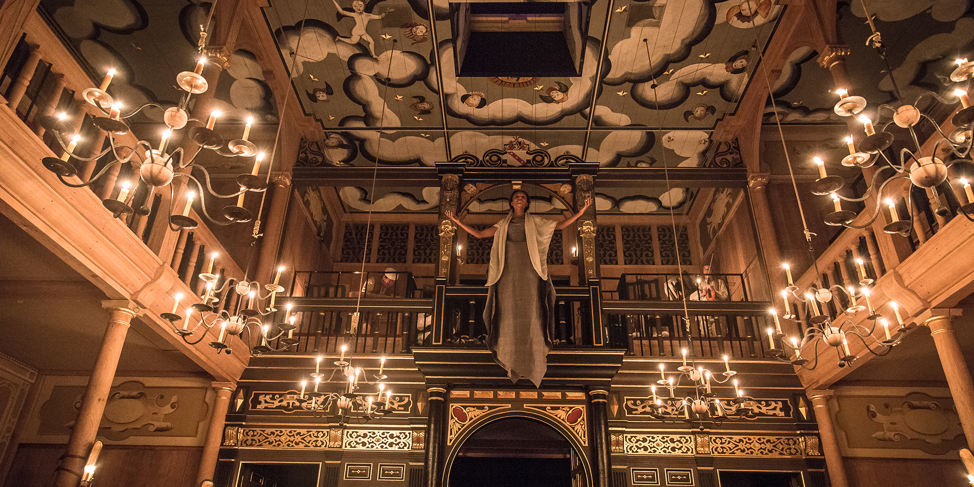 A woman descends from a trapdoor in the painted heavens of the Sam Wanamaker Playhouse, which is lit by candlelight