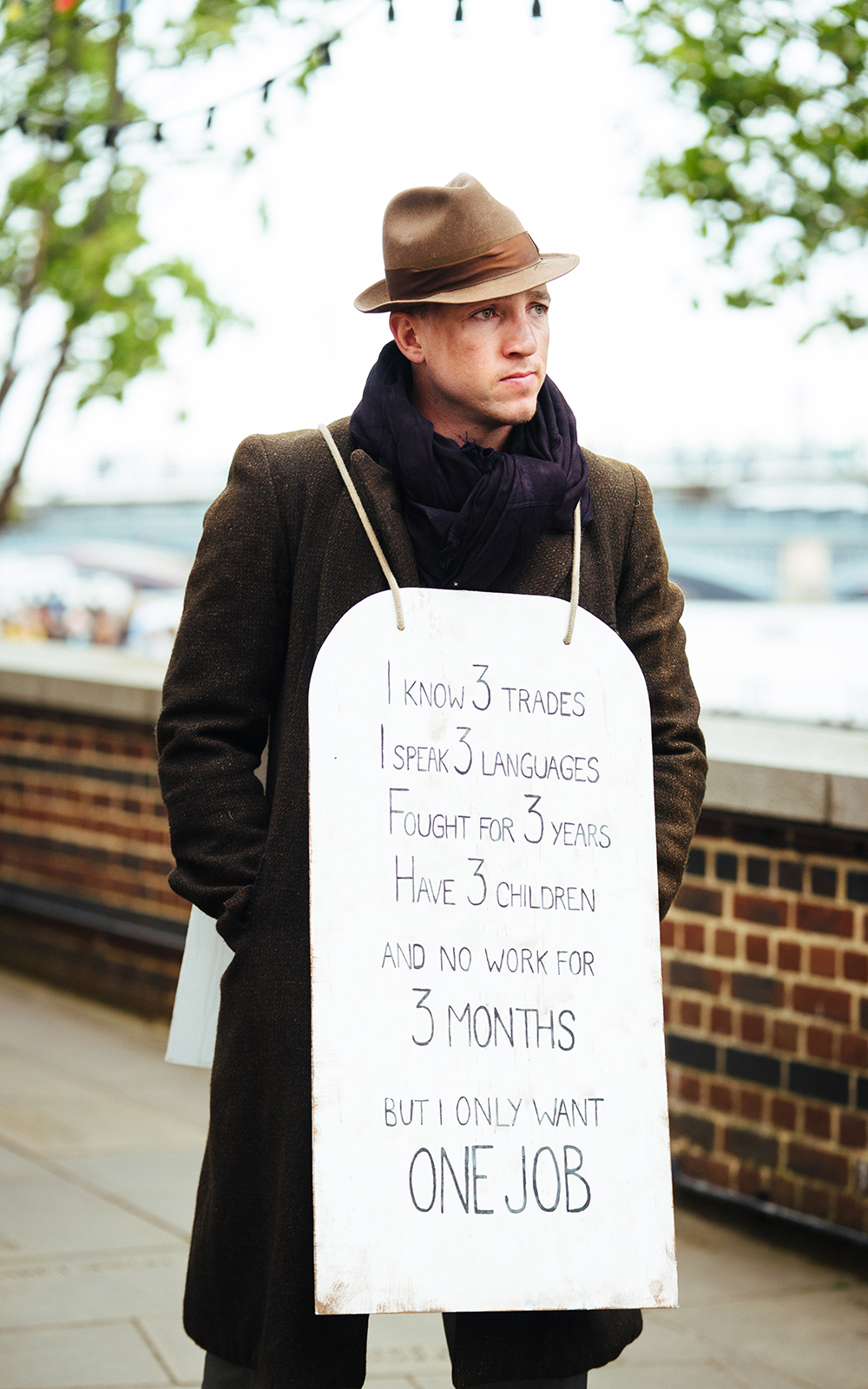 The Windsor Locals 4 Merry Wives of Windsor Shakespeare's Globe 2019 A man wearing a brown overcoat and hat stands glum with a billboard, outside.
