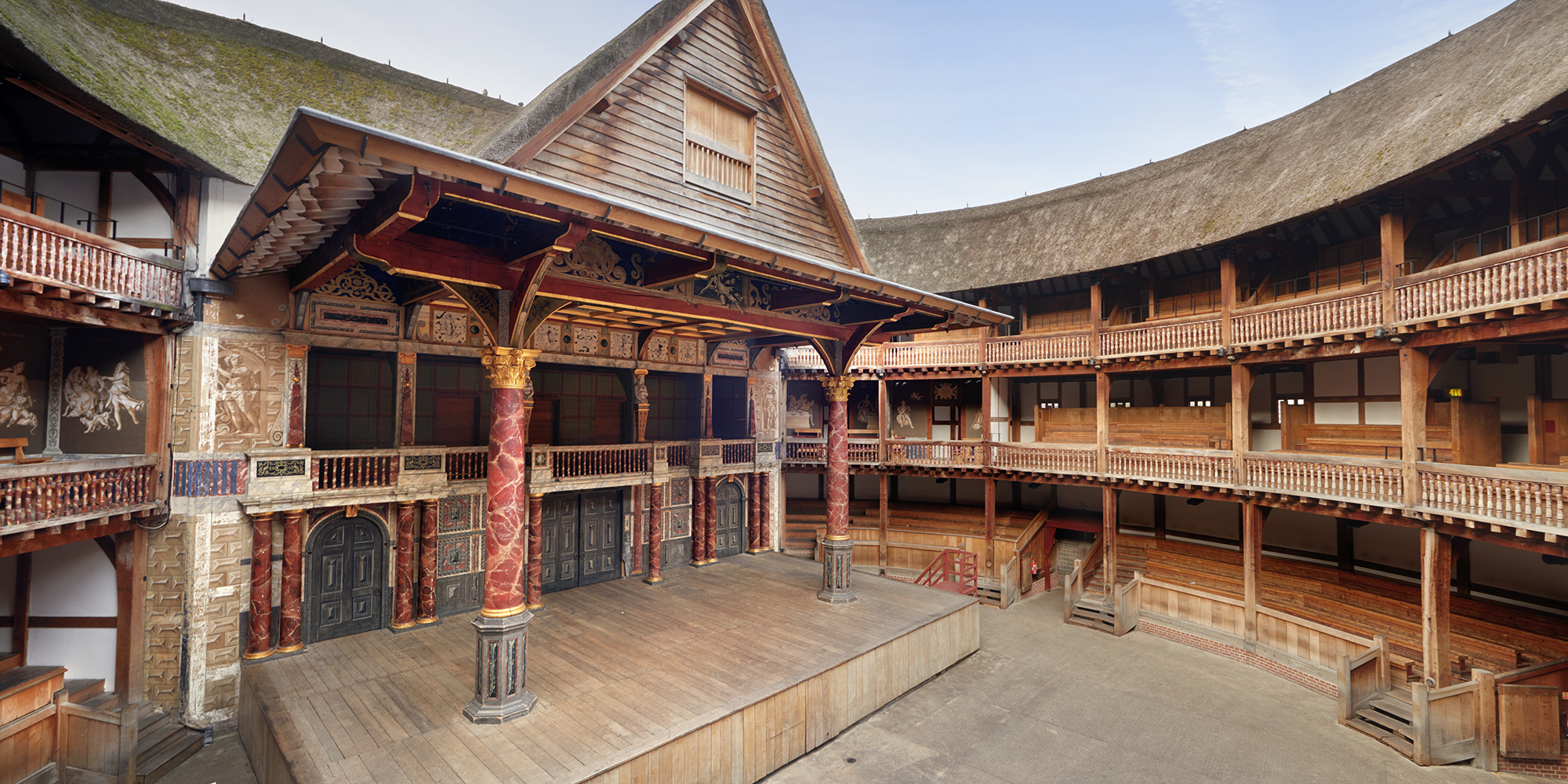 Interior shot of a circular timber theatre, with a wooden thrust stage, with two large pillars holding up a thatched roof