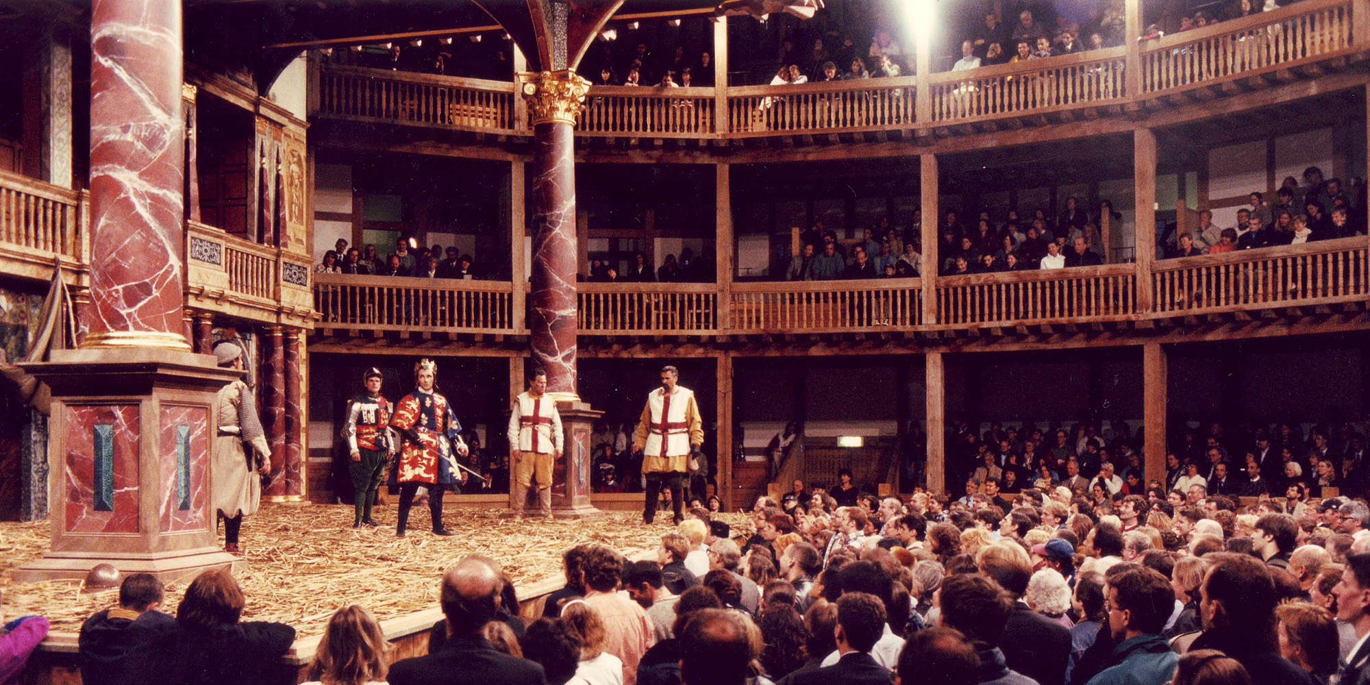 A large crowd of audience members stand watching a play in a circular timber structure.