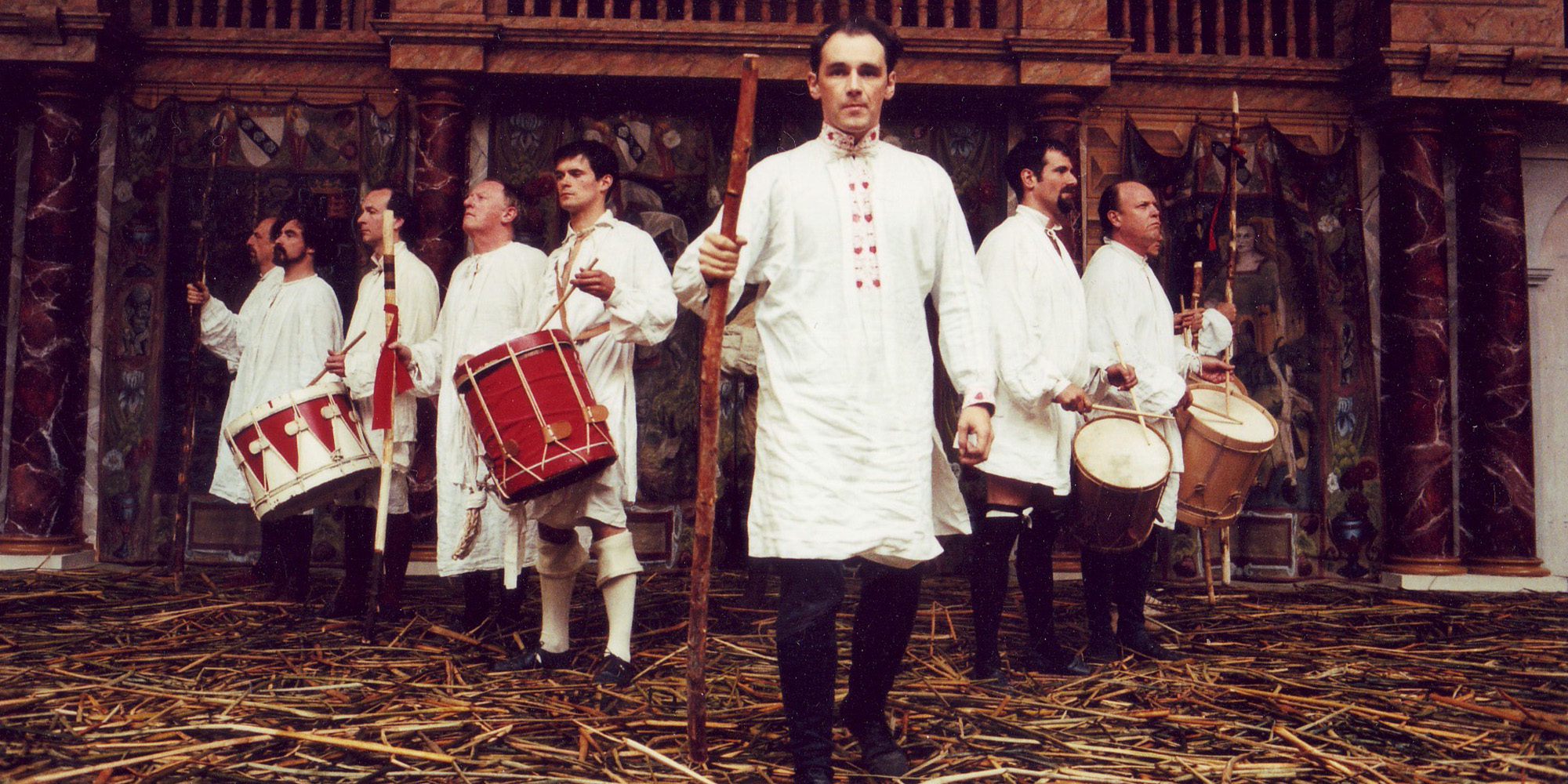 A group of actors wearing white tunics stand in a v-formation on a stage covered in straw.