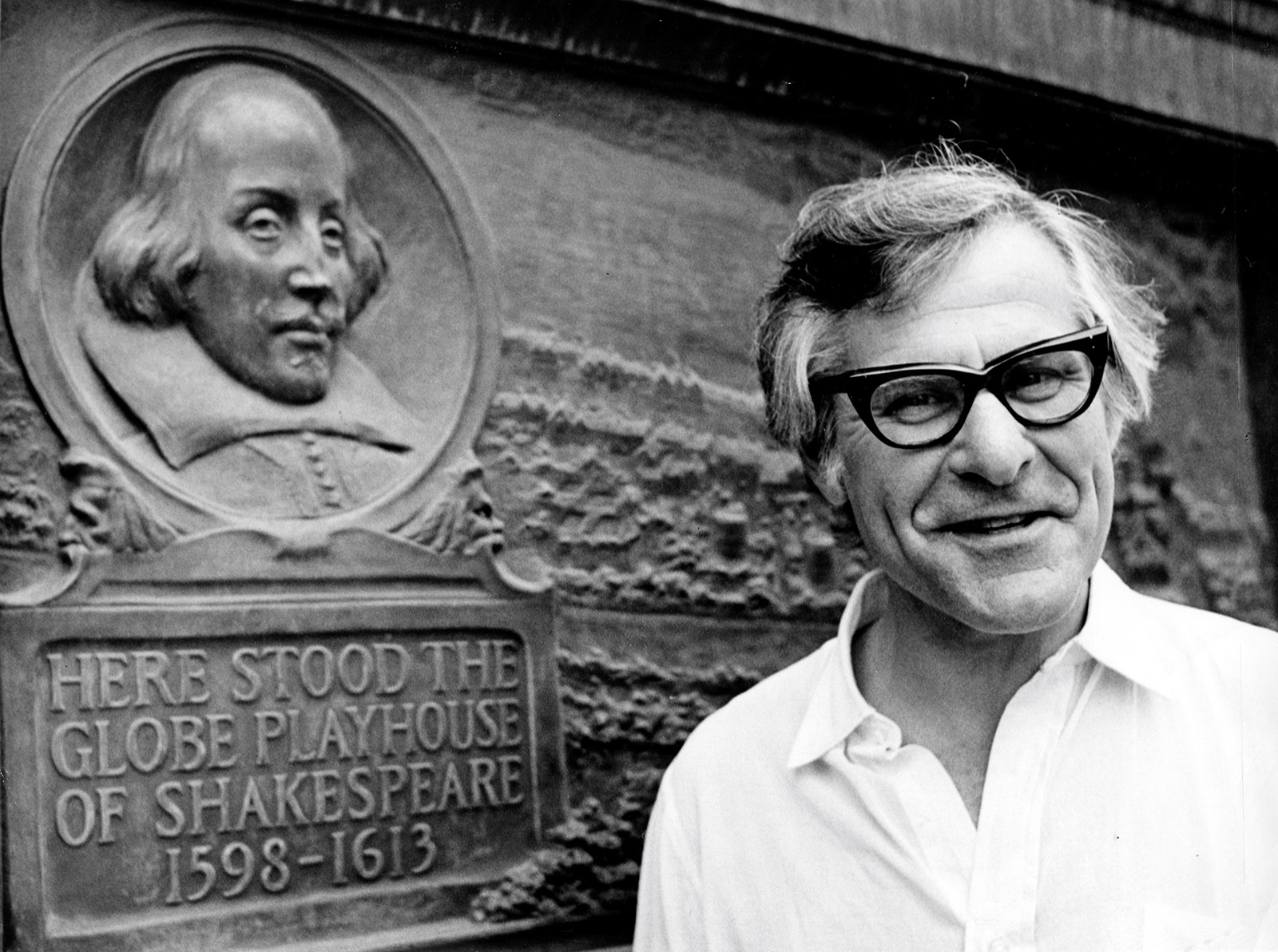 Sam Wanamaker Plaque Shakespeare's Globe A man wearing thick-framed glasses stands by a plaque marking the location of the original Globe Theatre