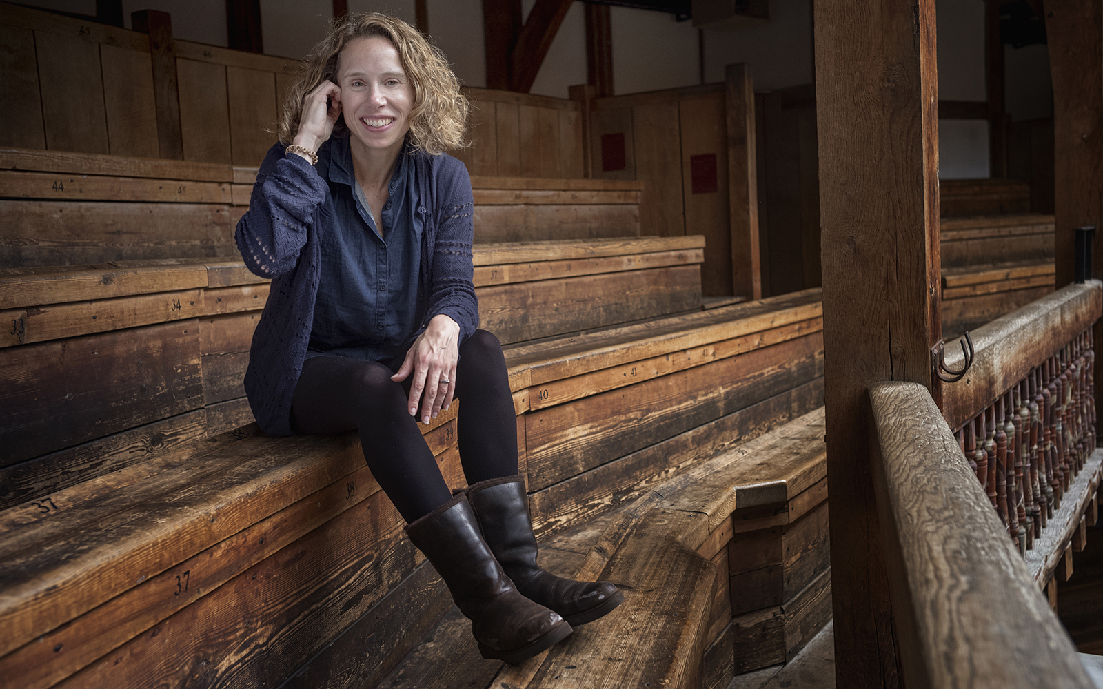 Globe Magazine Article Michelle Terry Artistic Director A woman sits smiling on a row of wooden benches, her hand tucking her hair behind her ear
