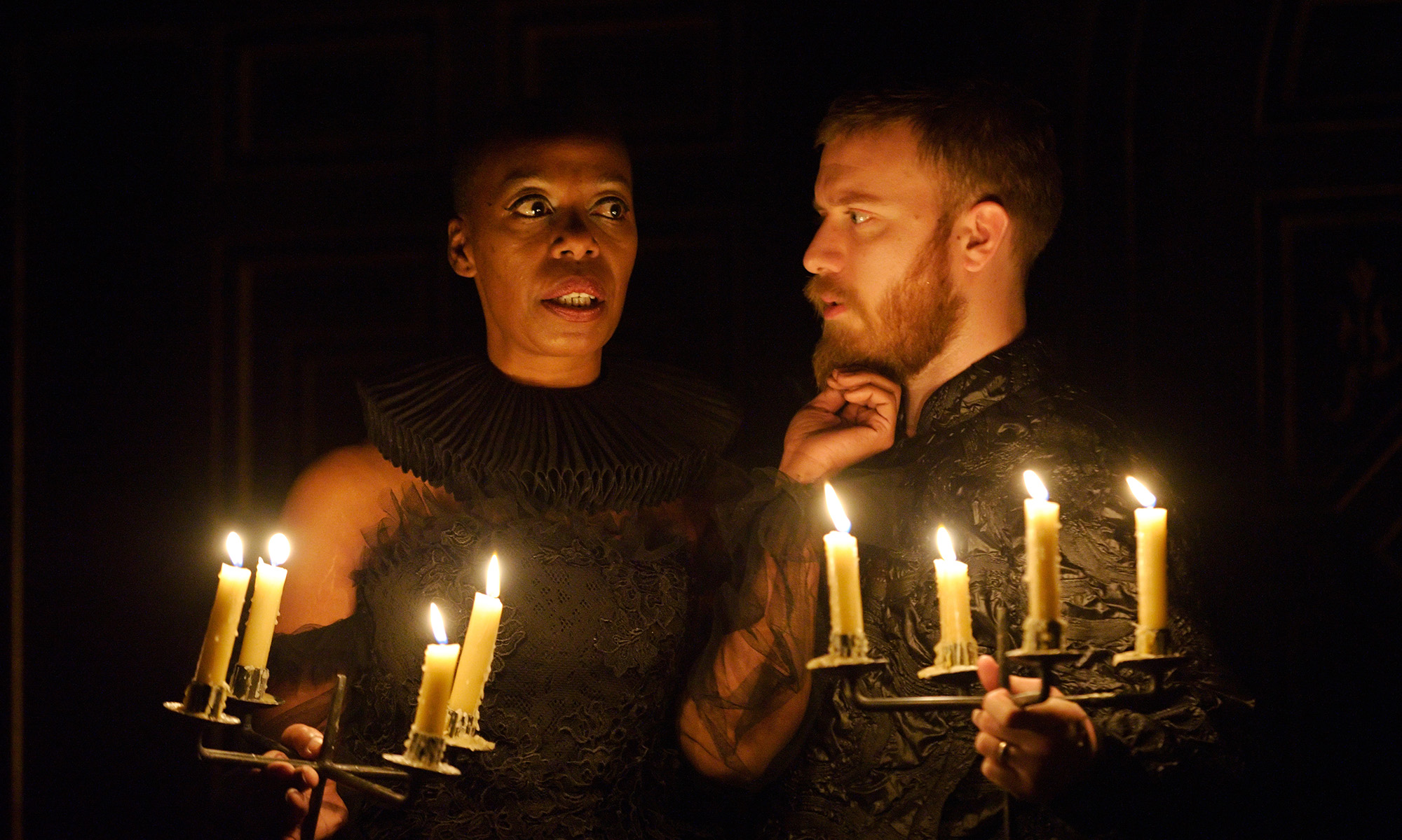 A woman and man stand in the glow of candlelight, the woman's hand touching the man's chin.