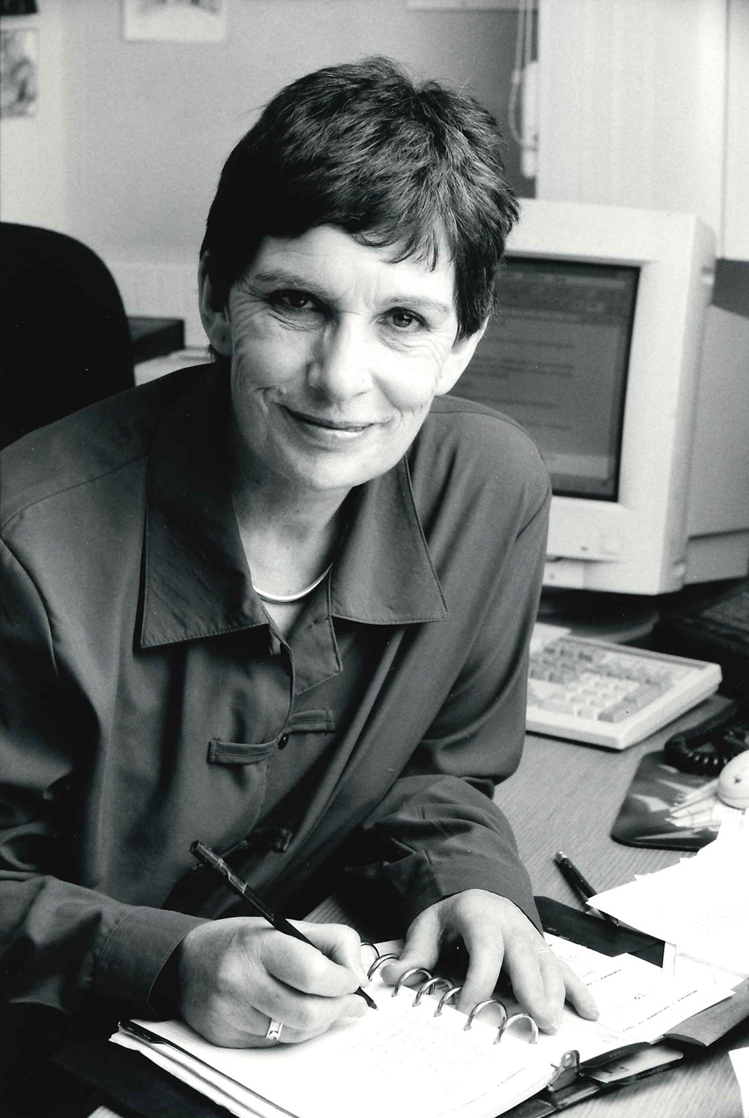 A black and white photograph of a woman smiling into the camera whilst writing in a notepad