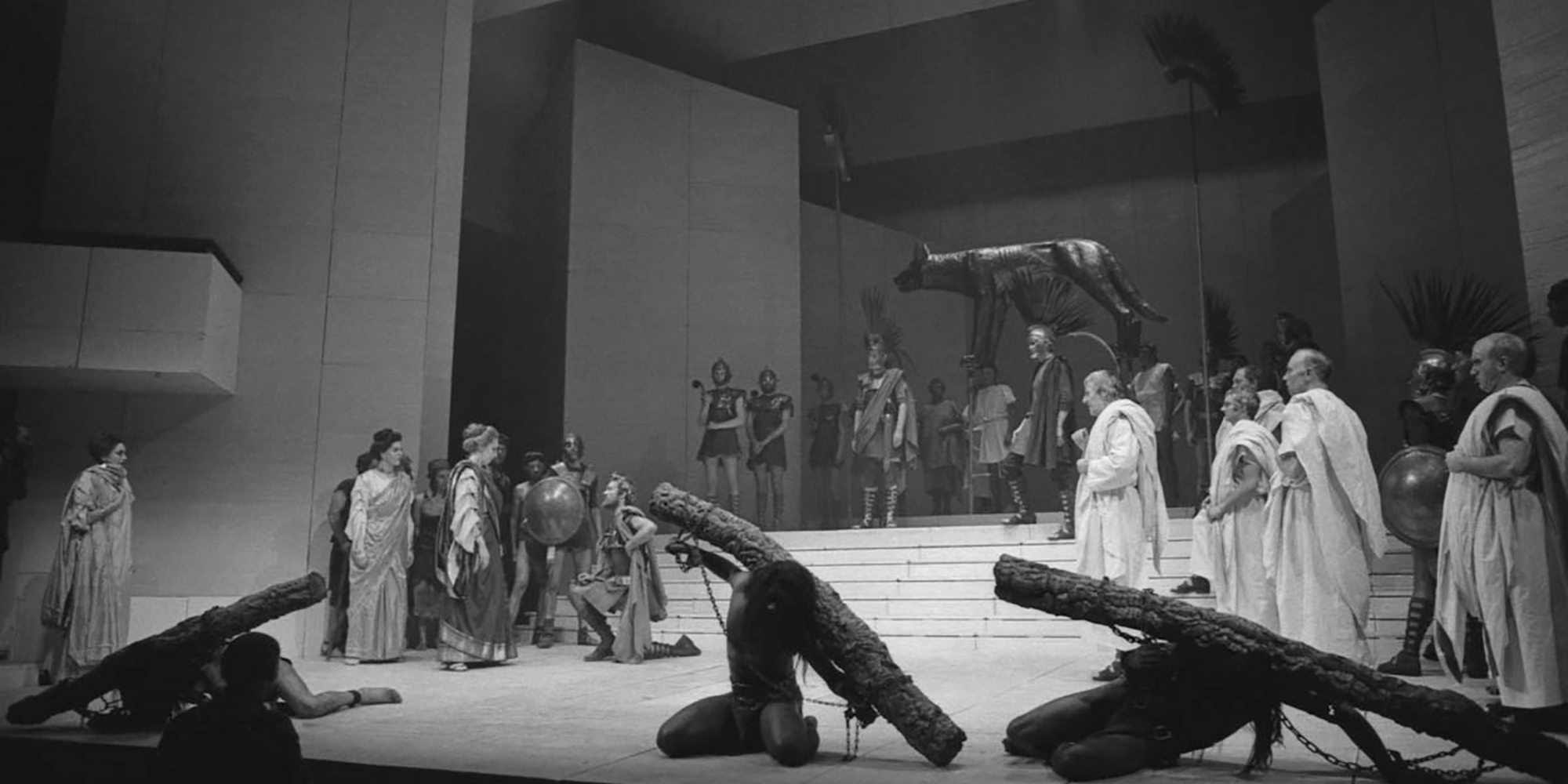 Black and white photograph of a proscenium stage, with a large group of actors standing in Roman garb: white tunics and robes, with plumed helmets.
