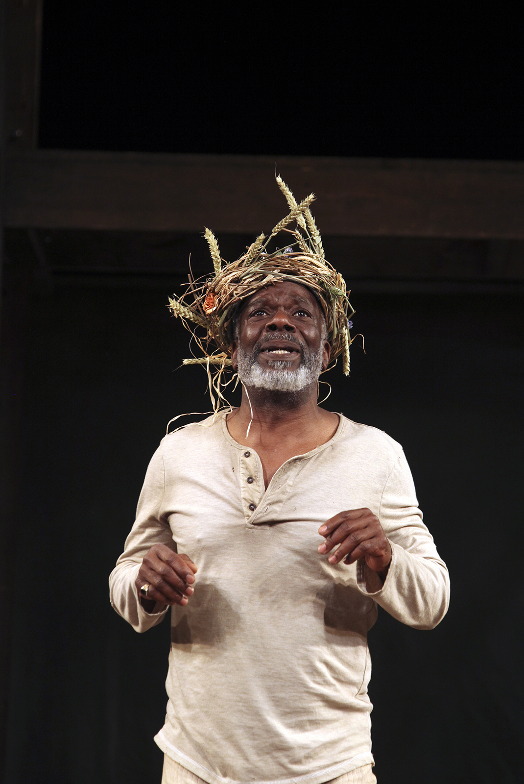 A man wearing white undergarments stands dismayed with a wreath of branches on his head.
