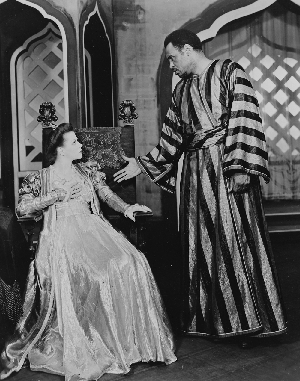 Paul Robeson Othello 1943-44 Library of Congress A black and white photograph of a woman sat in a chair, wearing an elaborate gown, with a man reaching out his hand next to her.