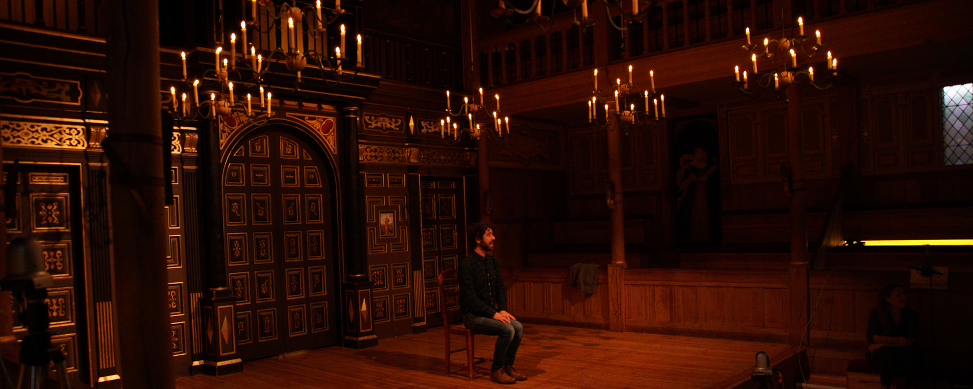 An actor sits on the stage surrounded by the candlelight of the indoor playhouse