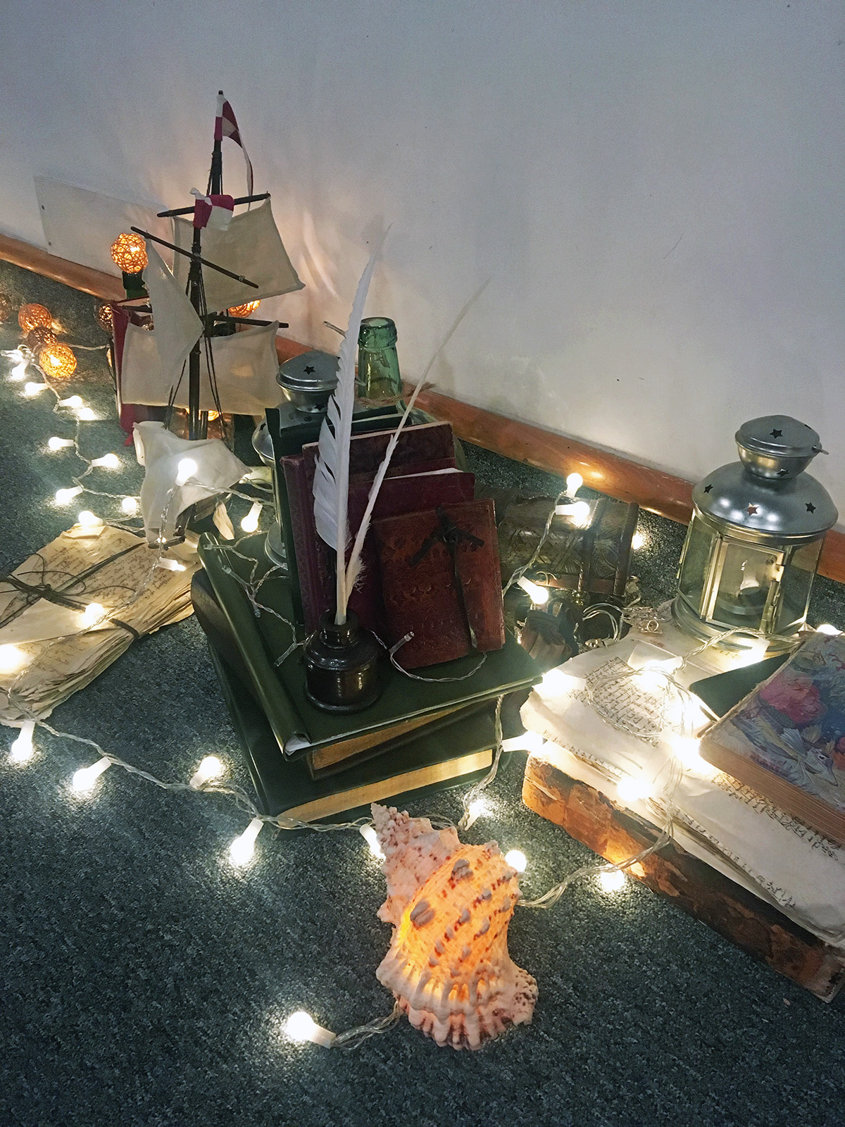 Fairy lights wrap around a pile of books, a model ship and lantern.