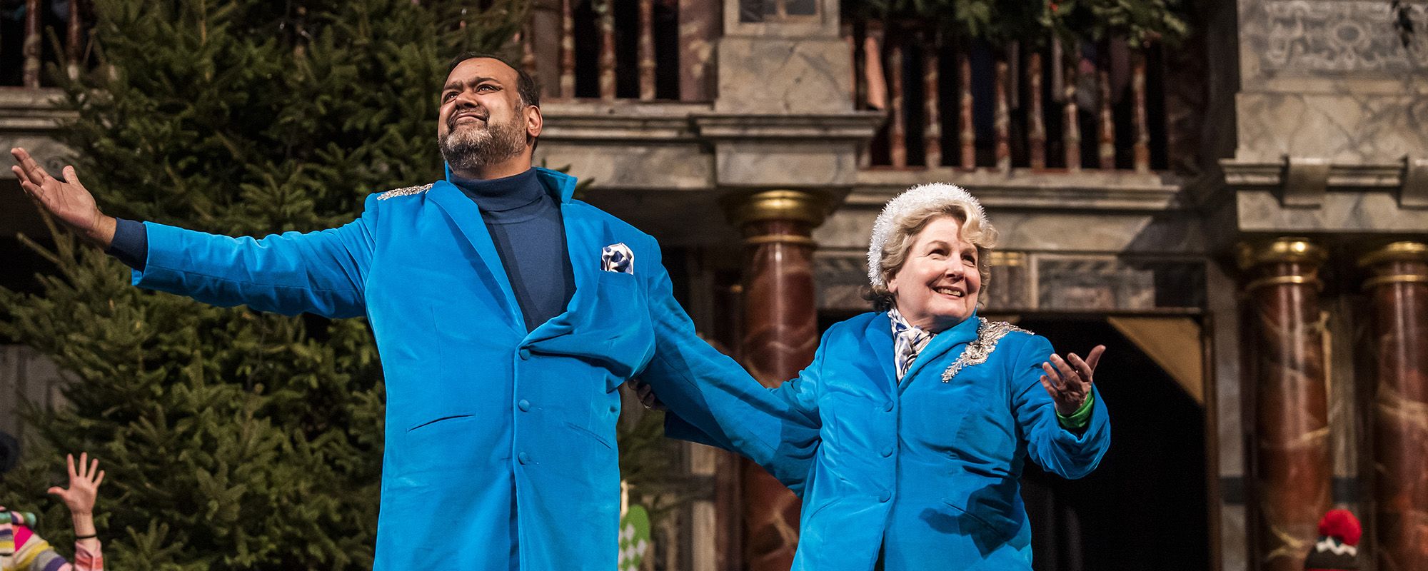 A male and female actor, wearing matching blue suits, stand onstage with their arms outstretched, smiling.