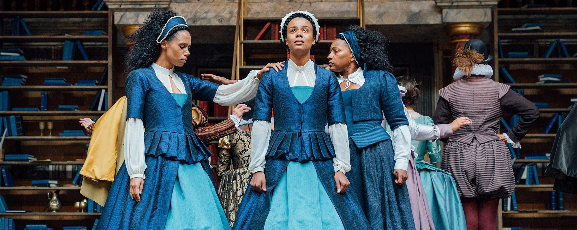 Emilia Shakespeare's Globe Helen Murray Three women, wearing similar blue Elizabethan dresses, stand together, each resting a hand on the other, supportive.