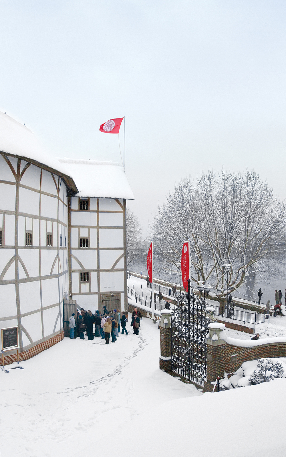 A snowy view of the white circular timber structure theatre, with the bank of the River Thames infront