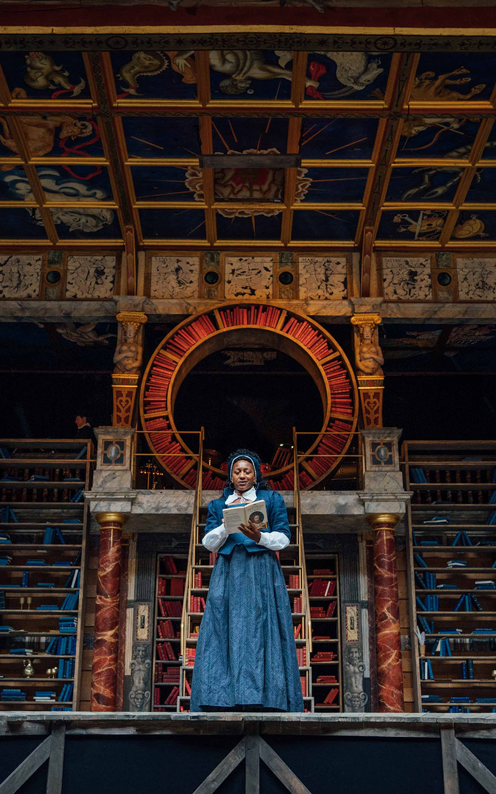 A woman wearing a blue Elizabethan corset dress stands on stage, reading a book, with shelves of bookcases behind her.