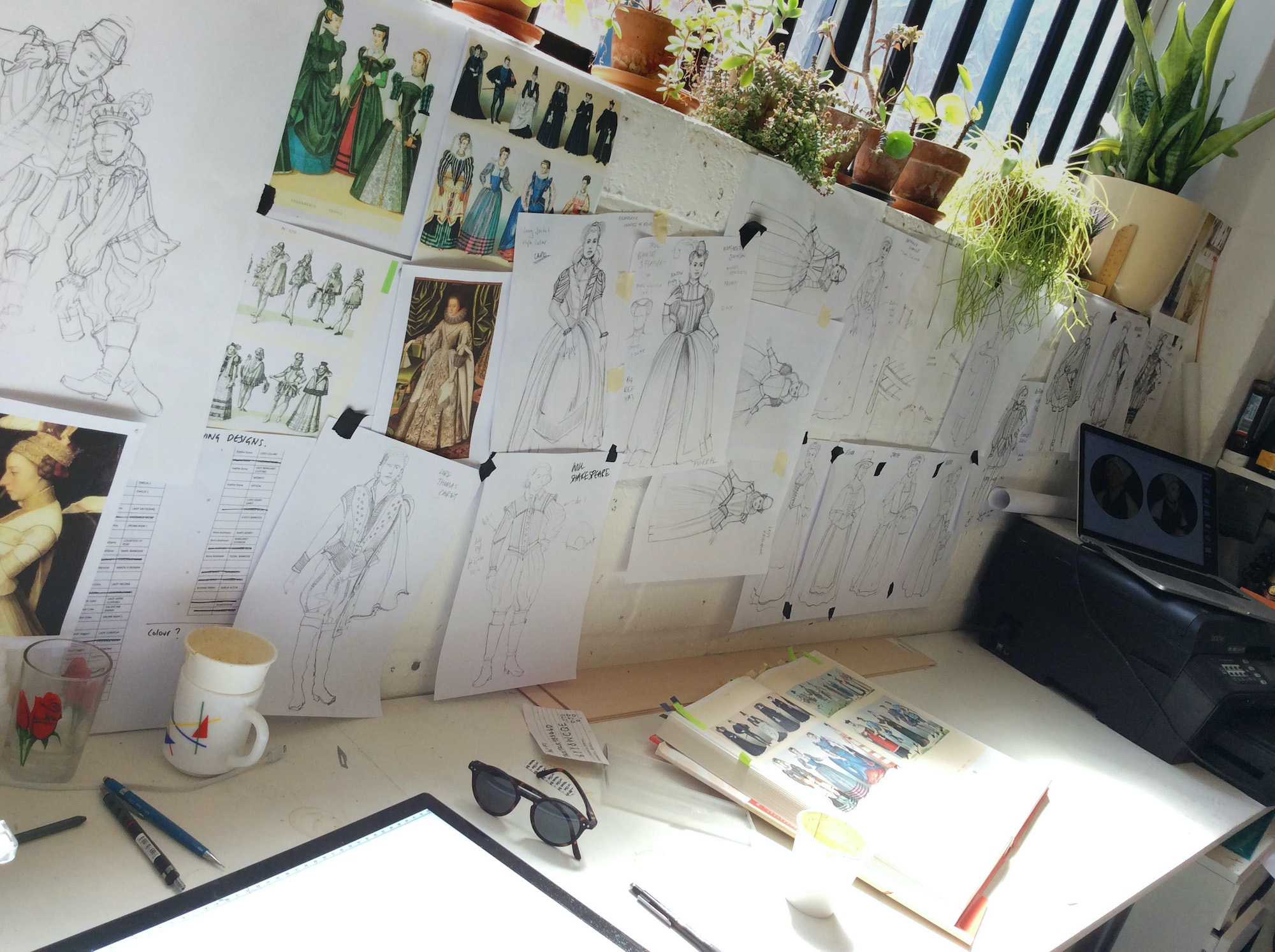 A photo of a designer’s desk shows sketches, a sketch book, a mug, a glass and pens