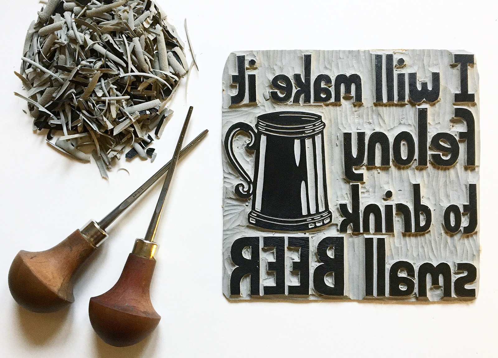 Carving a brand new card design Carving tools lay next to a block with a print of a beer tankard and the text: I will make it felony to drink small beer, in reverse