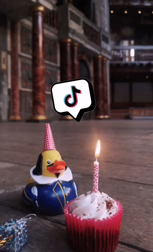 A rubber duck dressed as Shakespeare wears a red party hat and blows out a lit candle on a cupcake, with a TikTok speech bubble above his head.