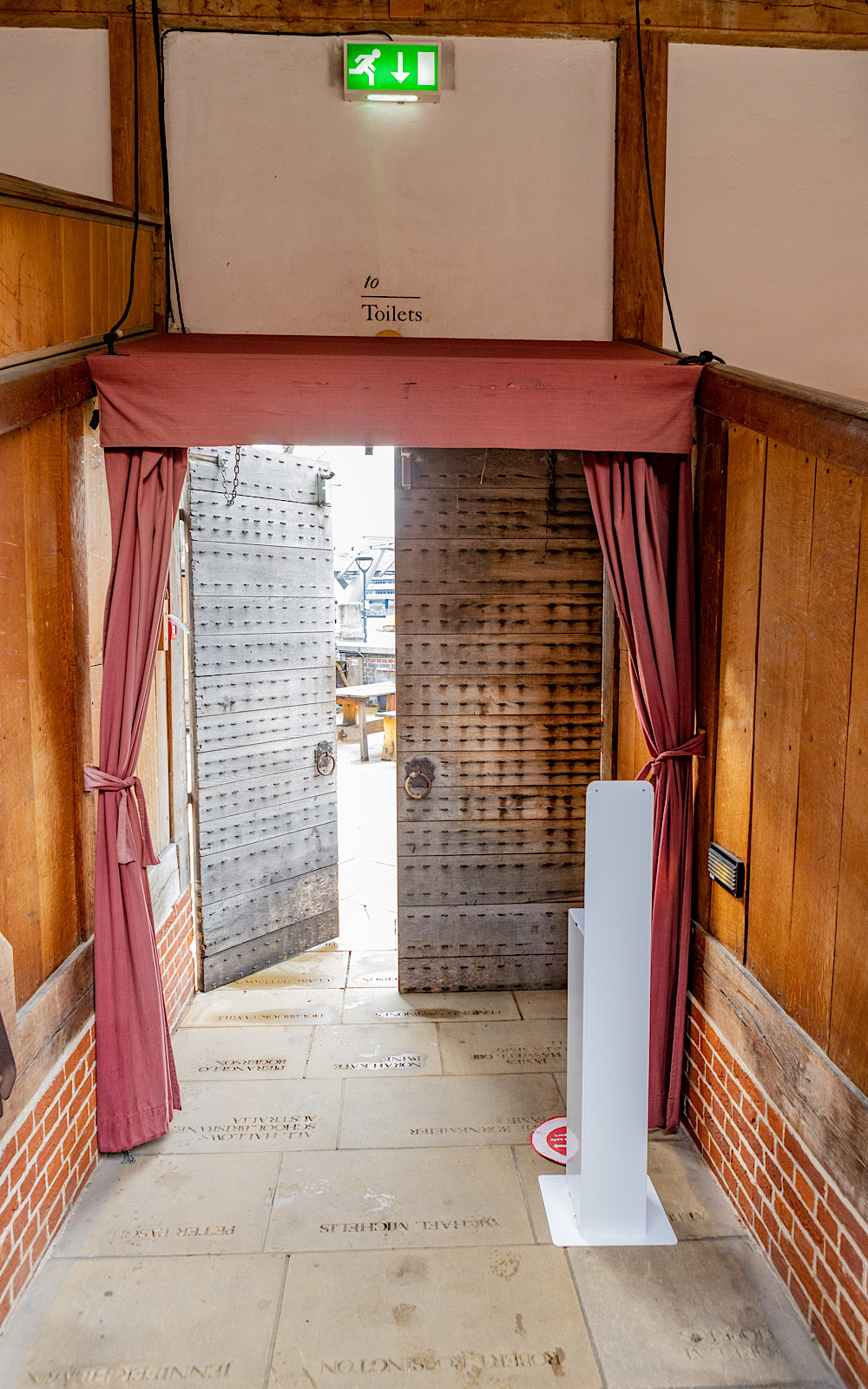 Covid Coronavirus Shakespeare’s Globe Pete Le May 2021 From the inside of a wooden theatre, doors lead to the outside, they have red curtains around them and a hand sanitising station behind them