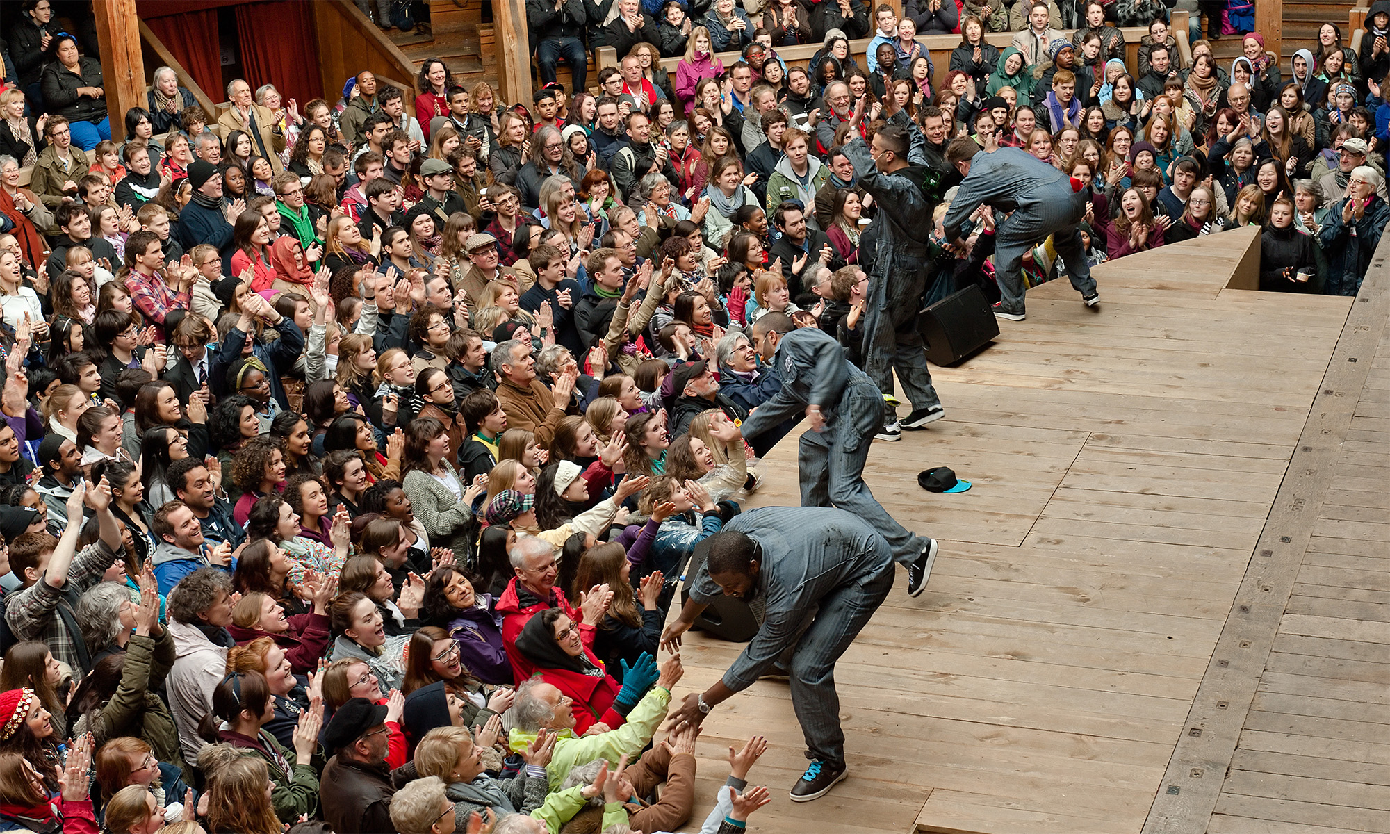 Four actors stand on the edge of a thrust stage, bending down to high five and hold the hands of audience members standing in the Yard in front.