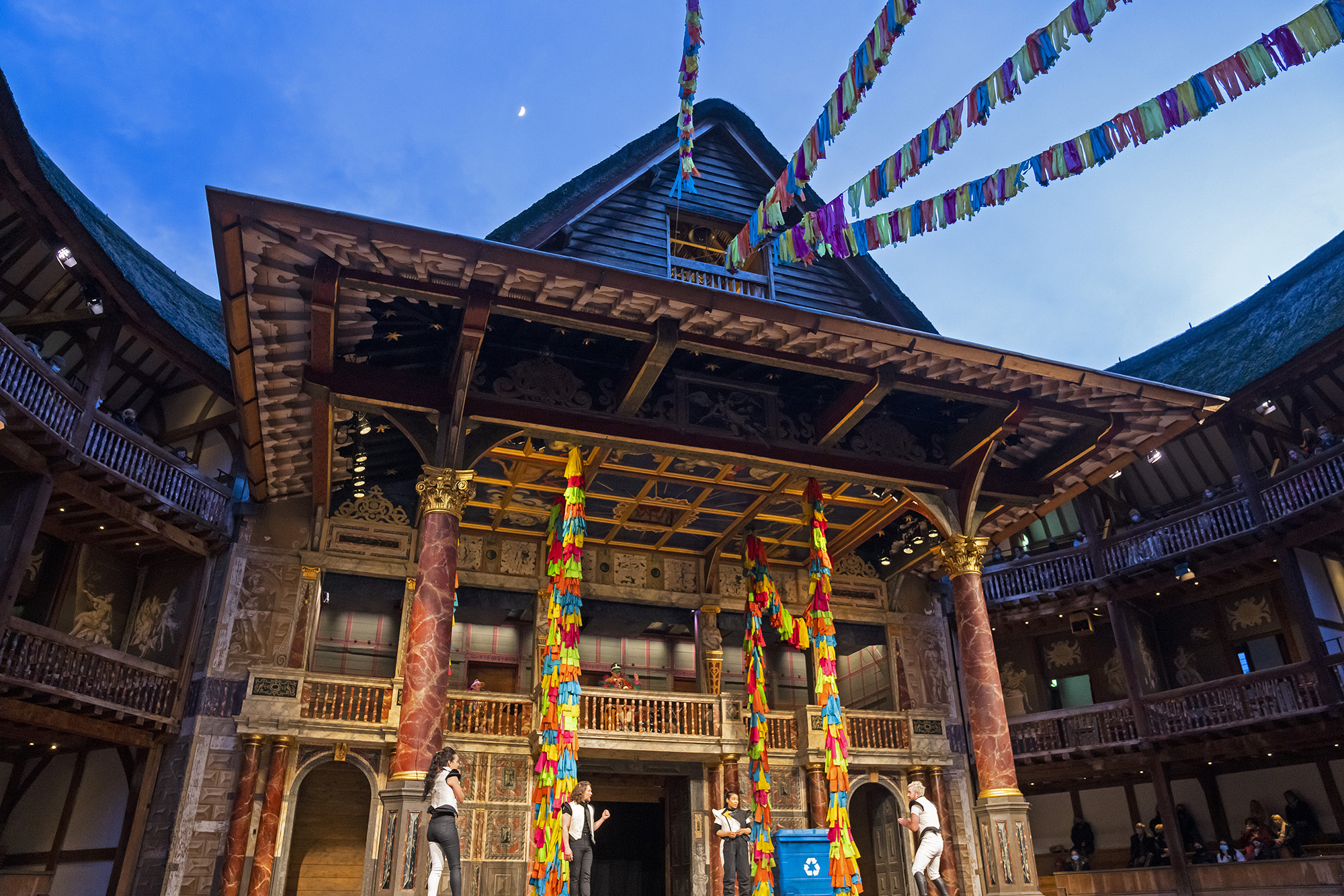 A twilight night sky, with a glimpse of the moon, shines above the Globe Theatre stage: with colourful streamings hanging across the open-air roof.