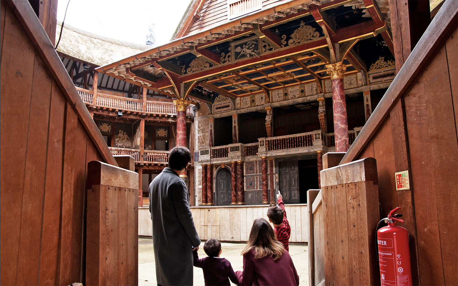 Two parents and two young children walk through the archway into the Yard of the Globe Theatre: pointing up at the painted heavens of the roof above the wooden stage.