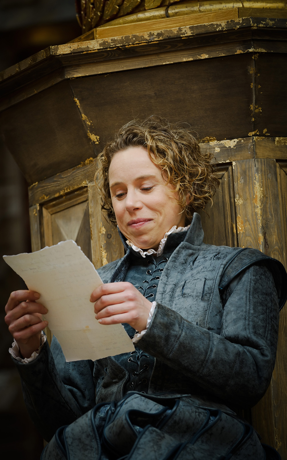 As You Like It An actor with short curly hair and wearing a doublet and hose sits reading a poem on a sheet of paper, smiling.