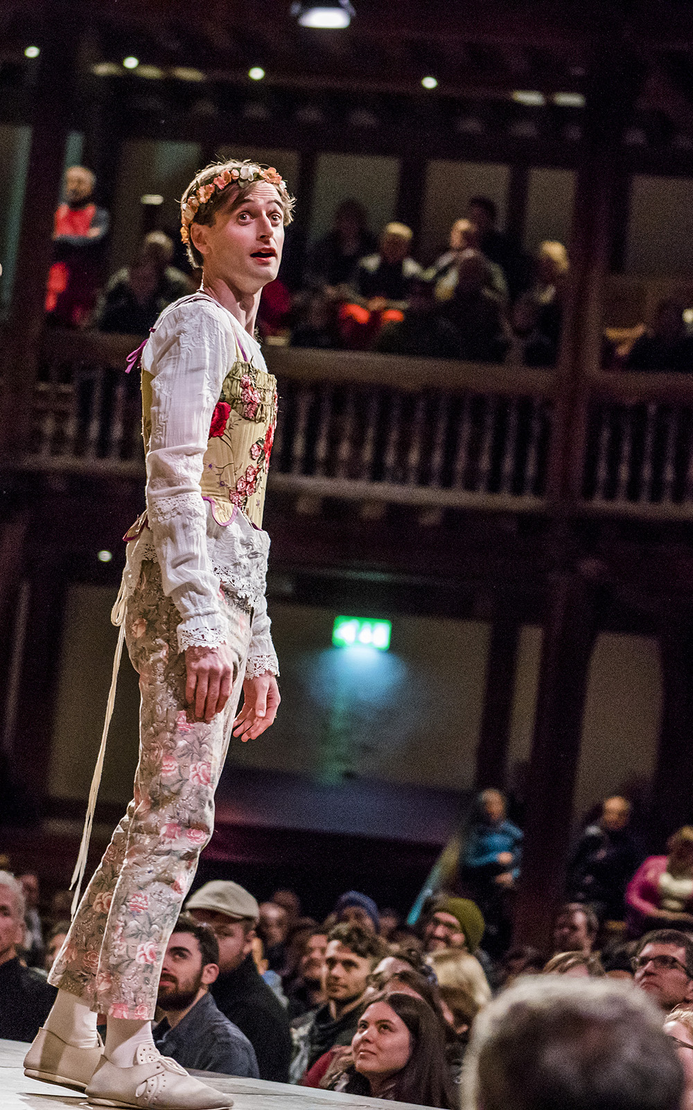 Who Is Rosalind As You Like It Shakespeare's Globe 2019 An actor wearing floral embroidered trousers and corset, over a white undershirt, looks over their shoulder, as audiences watch on.