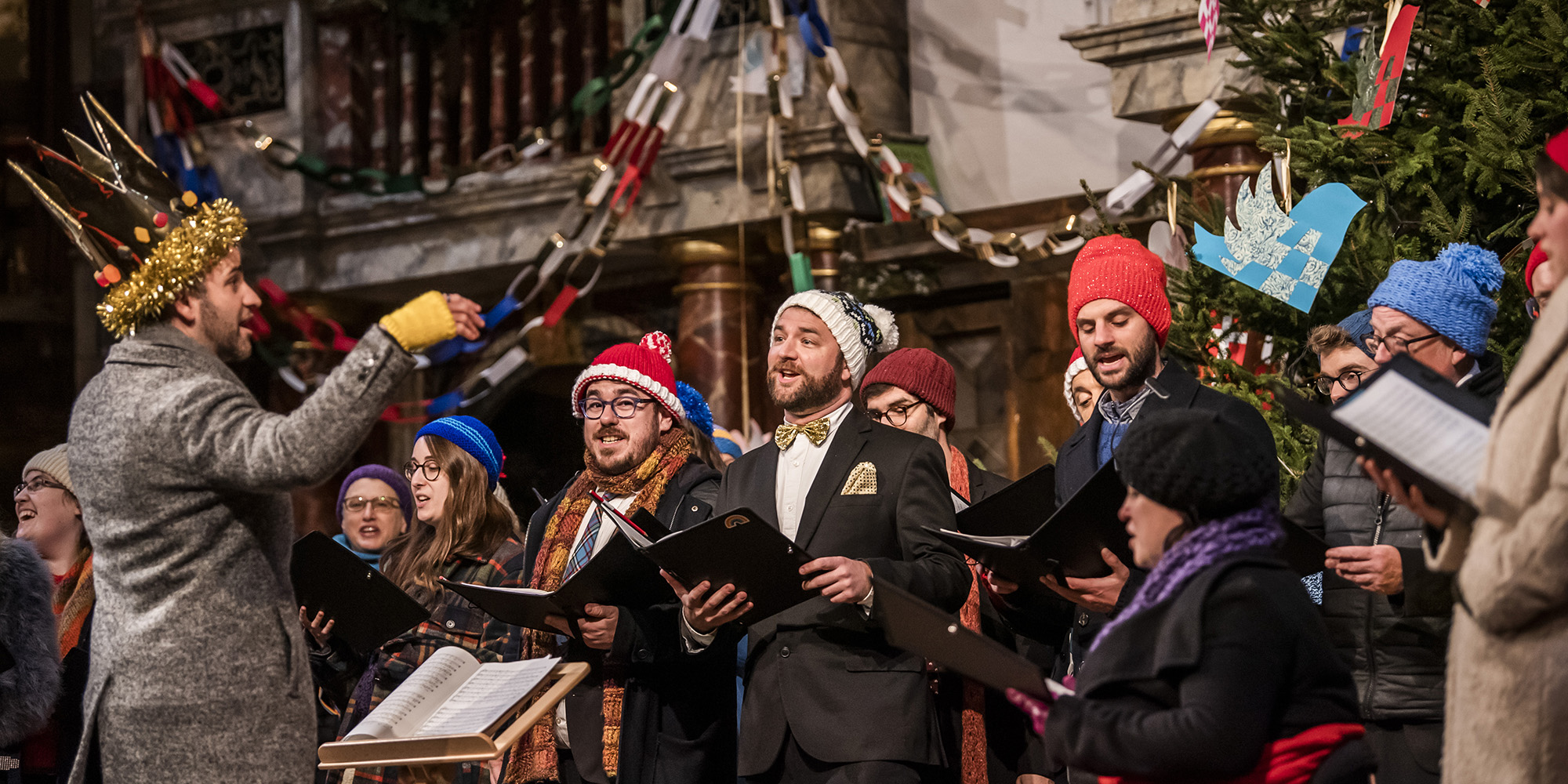 A group of choral singers wearing festive gear and Santa hats, stand on the Globe Theatre stage in front of a decorated Christmas tree, singing.