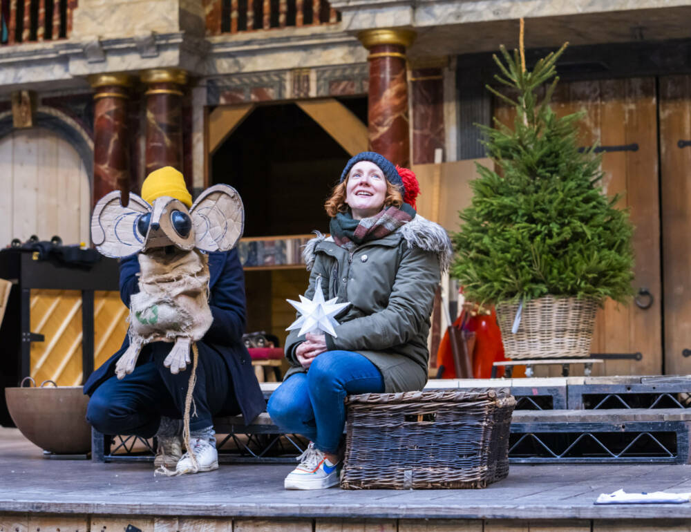 An actor sits on the Globe stage holding a white star, next to large cardboard puppet mouse. Behind them is a small fir tree.
