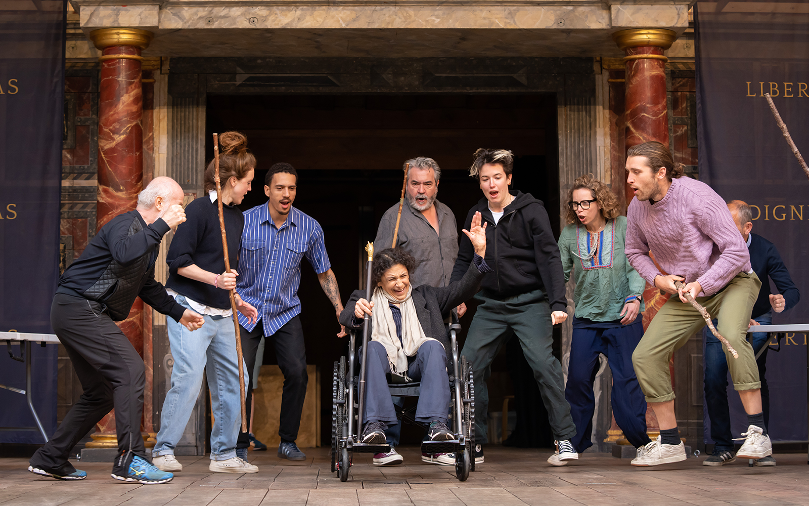A group of actors stand gathered around Kathryn Hunter in a wheelchair in the centre of the Globe stage.