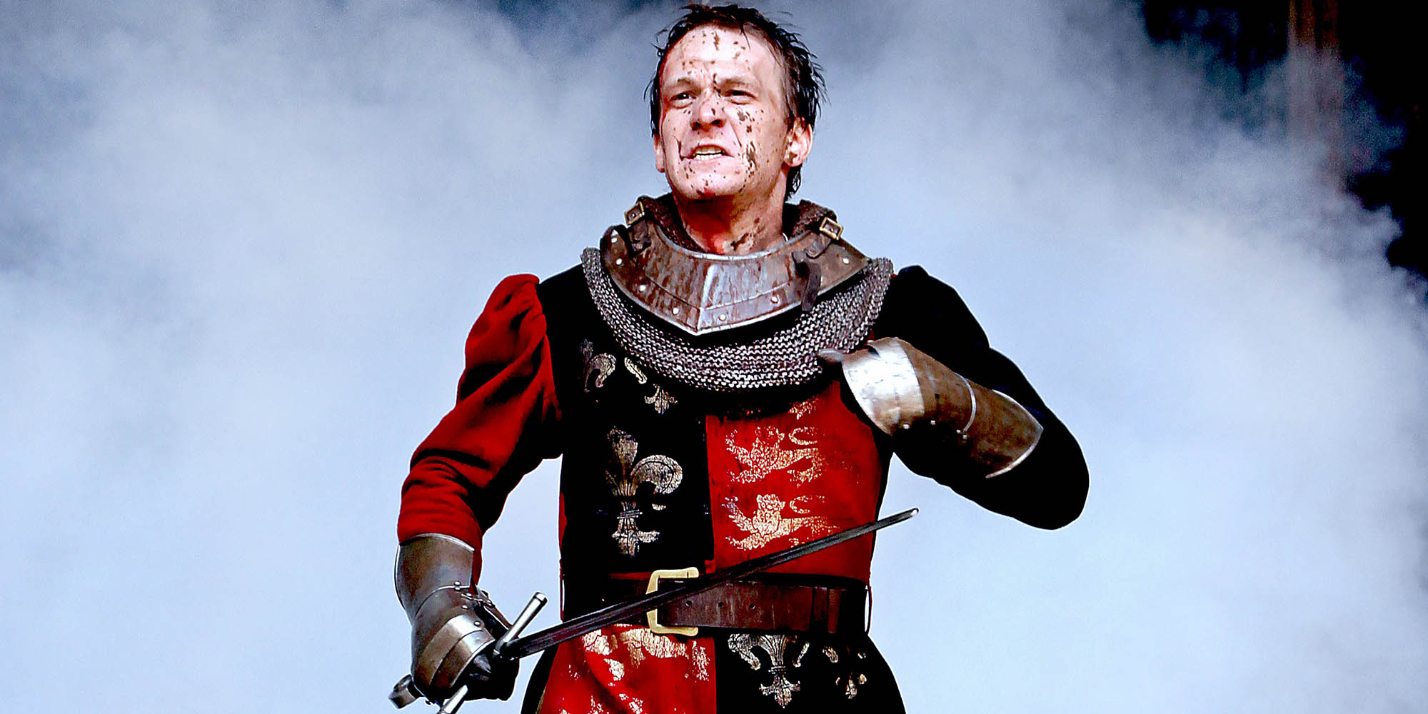 An actor wearing a traditional red and black English medieval military uniform runs holding a sword, with smoke billowing out behind them