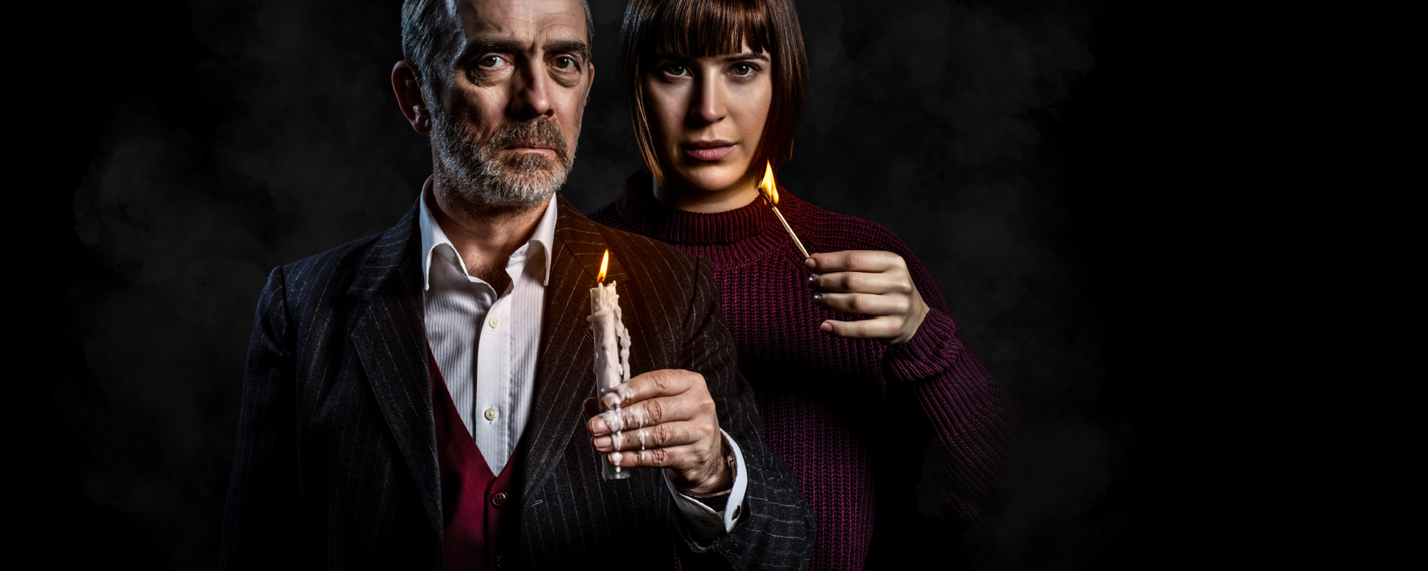 [IMAGE] Two actors stand close behind each other in the dark shadows. One is wearing a three piece suit, and holding a lit tapered candle that drips wax over their hand. The other is wearing a purple knitted jumper, and holds a lit match.