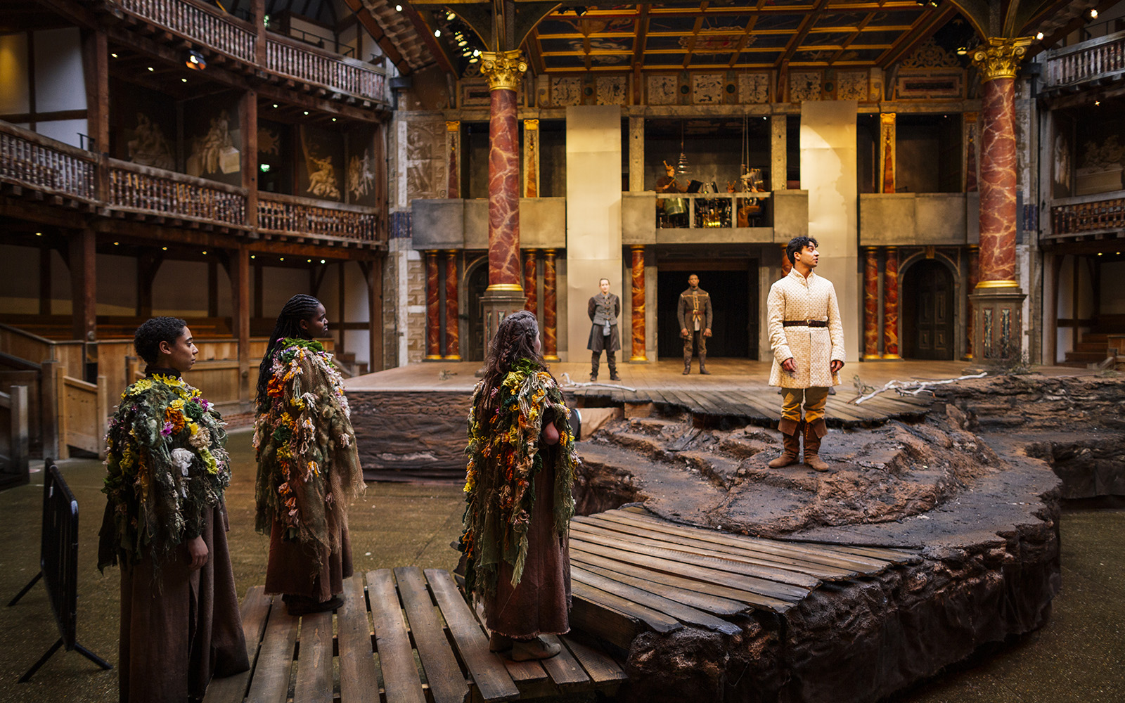 Three actors stand to the left of the stage adorned with flowers. Another actor stands in front of them dressed in a white tunic.