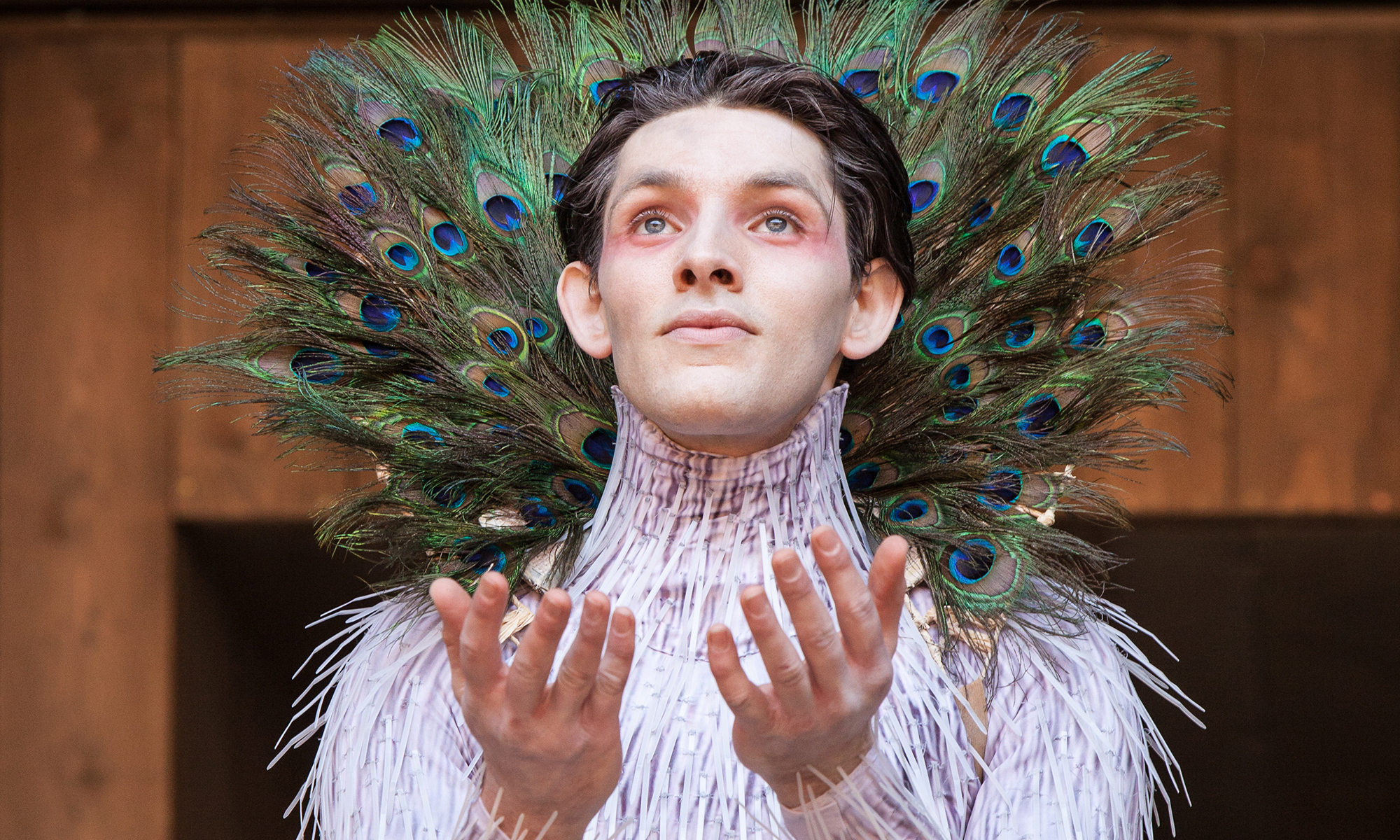 Colin Morgan Globe Player The Tempest Shakespeare's Globe flush An actor wearing a pink feathered costume and eye makeup, with peacock feathers fanned out in a plumage around their neck, holds out their hands palms upwards, looking up.