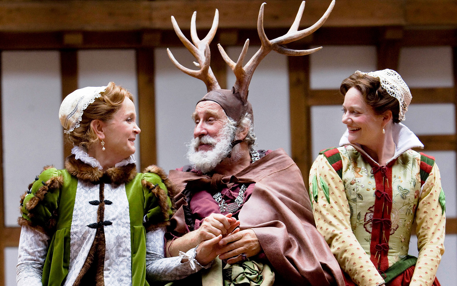 A man with a bushy white beard and antlers atop his head, sits inbetween to women wearing traditional Elizabethan dresses, who are laughing and smiling.