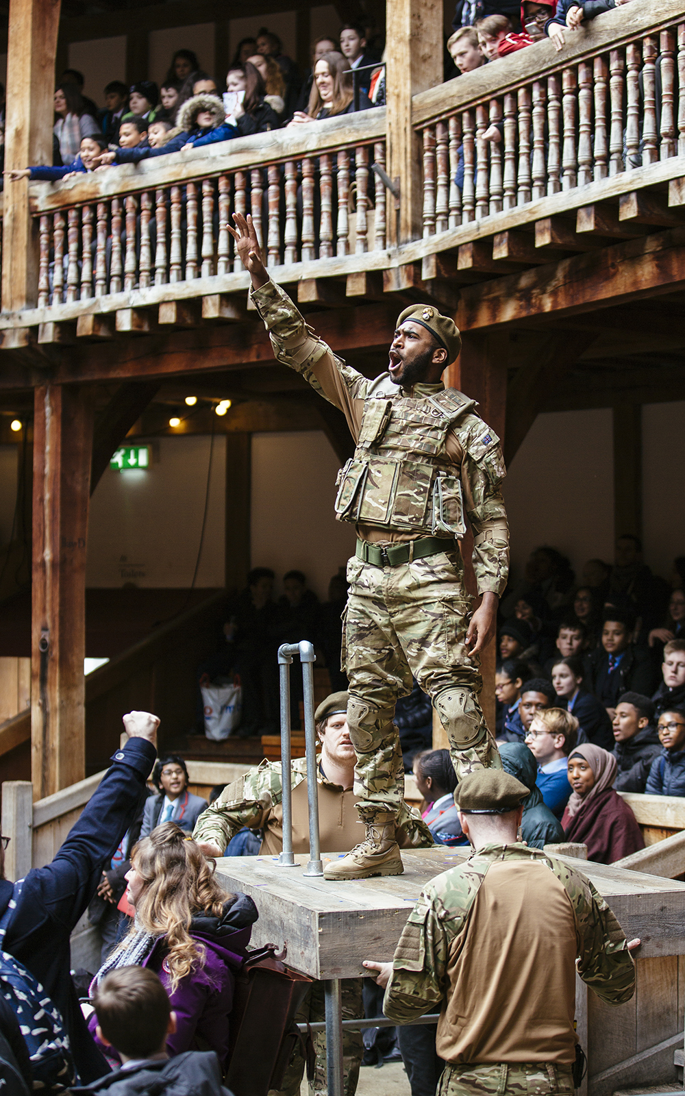An actor dressed in modern military cameo uniform stands on a plinth in the Yard of the Globe Theatre, their arm outstretched pointing to the sky and shouting.