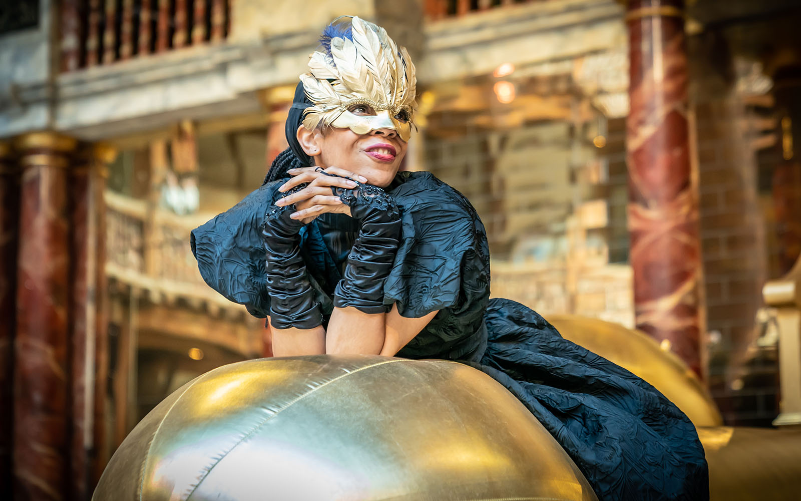 An actor wearing black corset dress, and golden feathered eye mask leans on a golden inflatable, smiling.