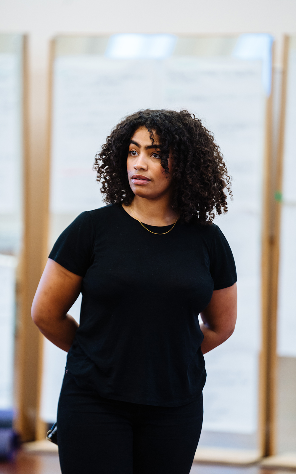 [image] a woman in a black t-shirt stands with her arms crossed behind her back, looking thoughtful to off-camera.
