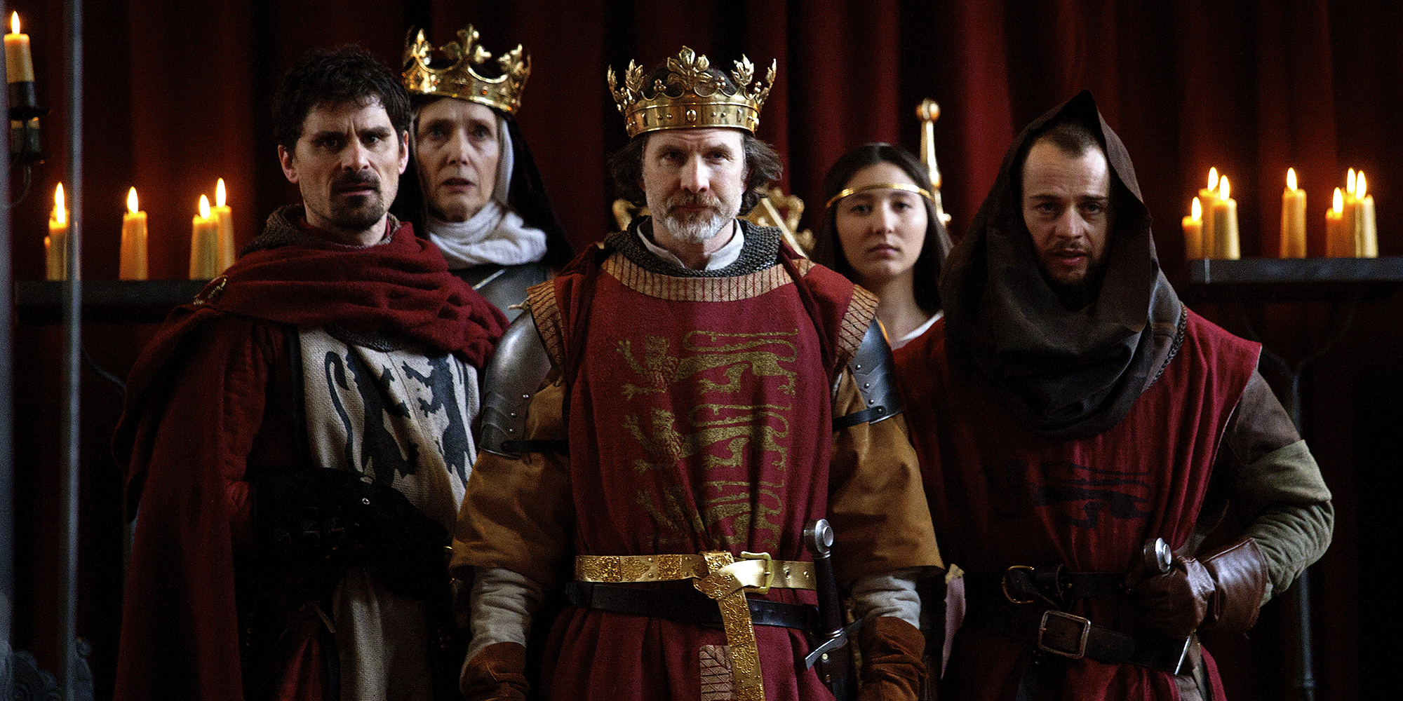 Five actors wearing medieval dress stand clustered together in a group. The actor to the centre wears a golden crown and red tabbard, embroidered with three golden lions.
