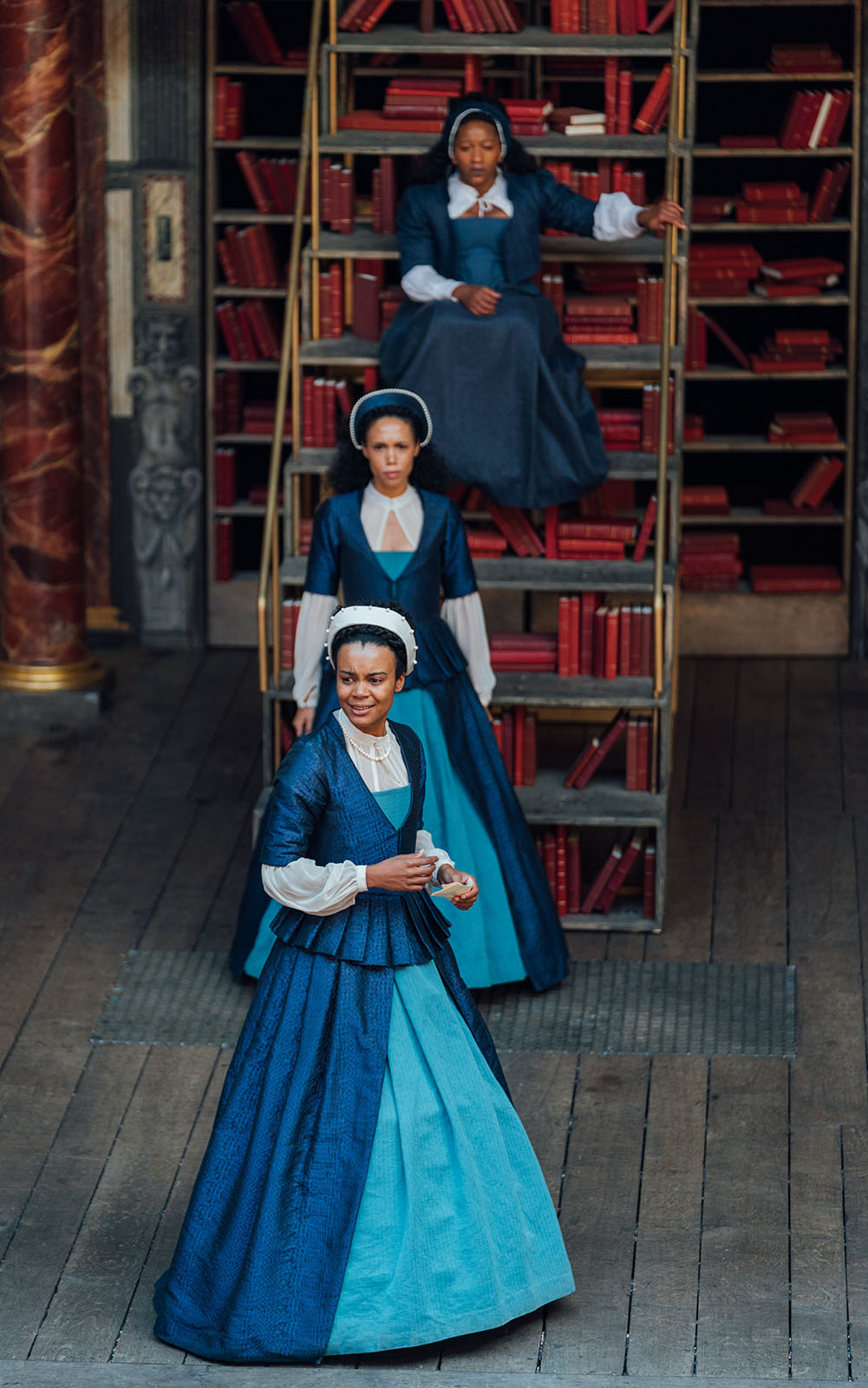 Three actors wearing blue Elizabethan corset gowns line up behind each other on the wooden floorboards of the Globe Theatre stage. The actor to the font holds their hands at their waist, smiling. The second actor stands directly behind, whist the third sits o a ladder staircase filled with red bound books.