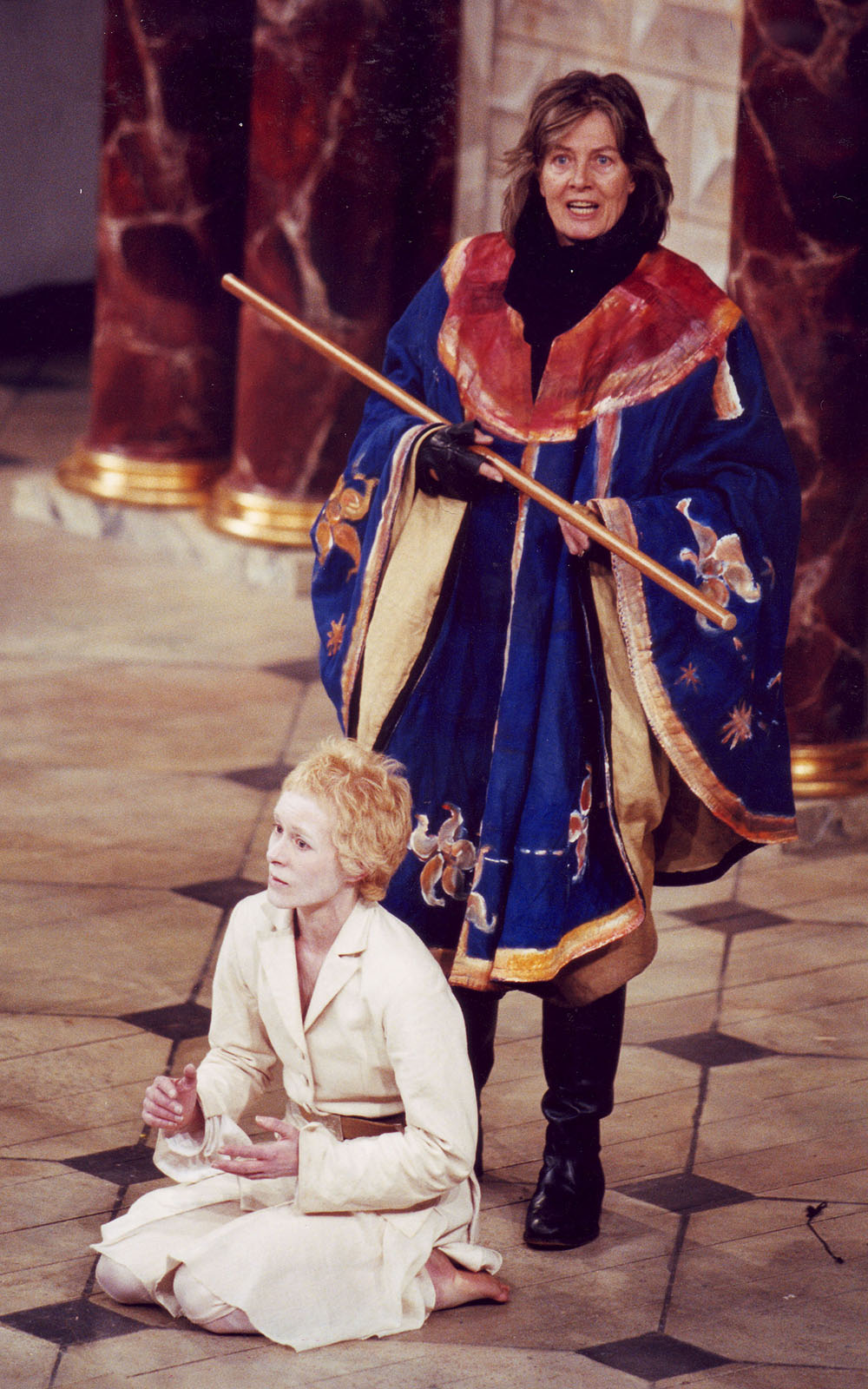 A woman in a cloak holding a staff stands over a pale-faced man dressed in white.