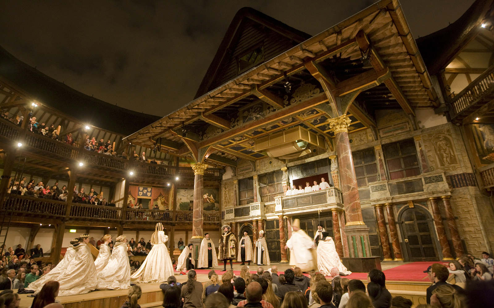 The Globe Theatre at night, cast in a golden glow. A procession of characters make their way up an extended catwalk to the Globe stage, as audiences watch on.