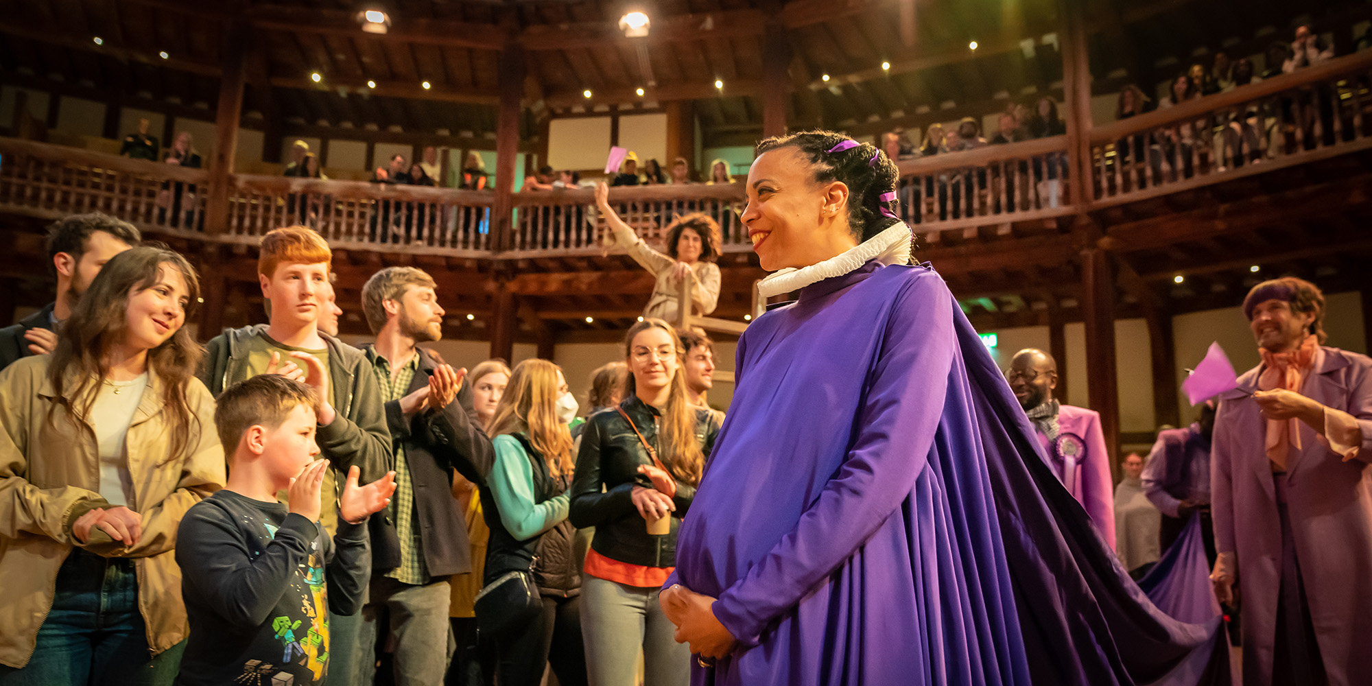 Henry VIII Shakespeare's Globe 2022 Marc Brenner flush An actor wearing a deep purple gown and ruff, covering a visible pregnancy bumps, beams as they walk through audience members applauding and waving purple flags in the Yard of the Globe Theatre.